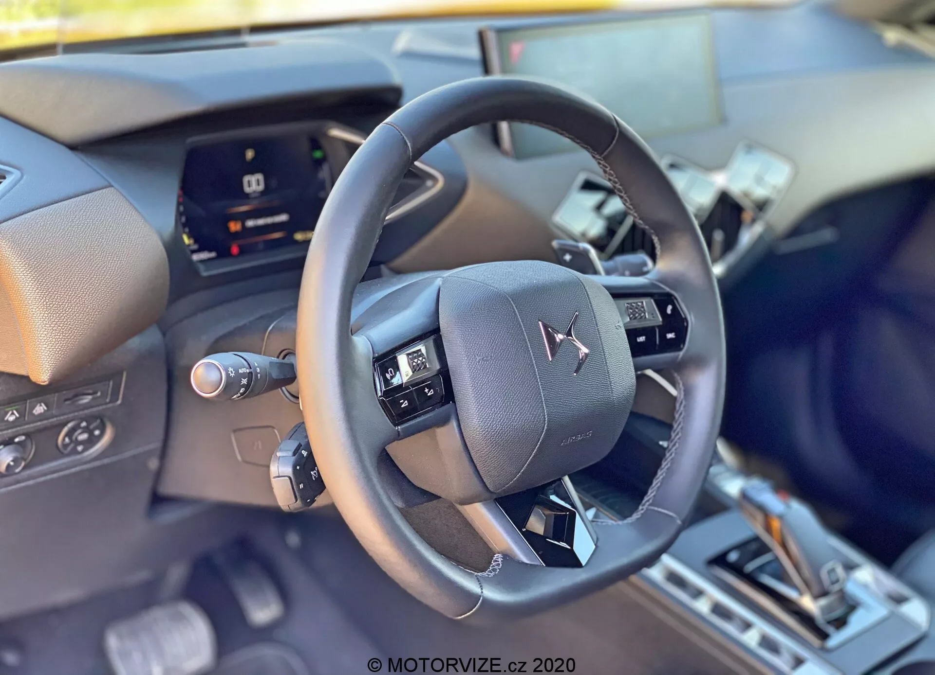Zbliżenie wnętrza DS Automobiles DS-3 2019 (DS-3 Crossback) z perspektywy kierowcy, skupiające się na nowoczesnej skórzanej kierownicy znajdującej się w centrum kadru z wielofunkcyjnymi kontrolkami. Za kierownicą znajduje się częściowo zasłonięty cyfrowy zestaw wskaźników, wskazując zaawansowaną technologię pojazdu. Dolna część obrazu sugeruje konsolę centralną z dźwignią zmiany biegów i możliwymi przełącznikami trybów jazdy, ukazując wyszukane wnętrze i cechy komfortu.