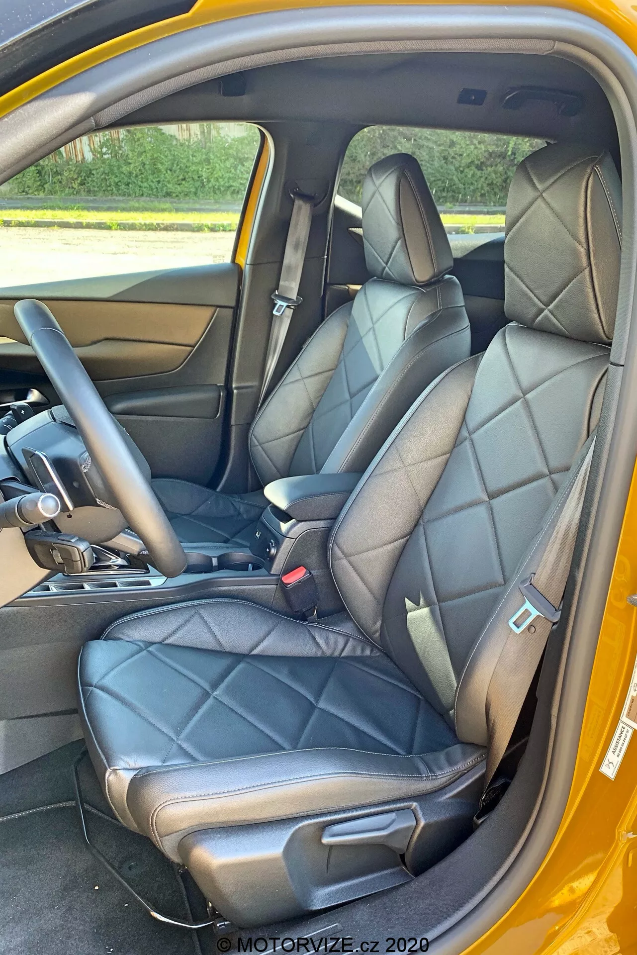 Widok wnętrza DS Automobiles DS-3 Crossback 2019 skupiający się na przednich siedzeniach z perspektywy drzwi pasażera. Obraz podkreśla czarne skórzane pikowane fotele z zintegrowanymi zagłówkami i bocznymi podparciami, sugerując luksusowy lub sportowy design. Widoczna jest część środkowej konsoli z dźwignią zmiany biegów wskazującą na automatyczną skrzynię biegów oraz dolna część deski rozdzielczej. Panel wewnętrzny drzwi odpowiada skórze siedzeń, z metalową klamką i przyciskami sterowania. Widać również czarne pasy bezpieczeństwa z mechanizmem regulacji wysokości, wykładzinę podłogową i kontrastujące zewnętrzne schemat kolorów, możliwie żółte.