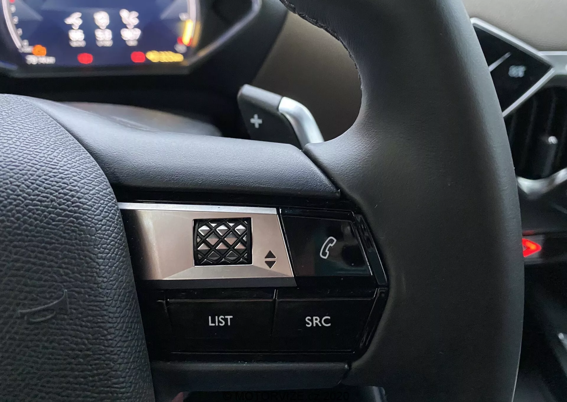 Bliskie ujęcie z perspektywy kierowcy na lewą stronę kierownicy w DS Automobiles DS 3 Crossback 2019. Obraz koncentruje się na przyciskach sterujących, w tym przycisku z ikoną telefonu, przycisku 'LIST' i przycisku 'SRC', podkreślonych na rozmytym tle deski rozdzielczej. Wysoki kontrast w projekcie łączy czarne i metaliczne wykończenia z wzorem diamentowym na chromie, podkreślając wygląd premium.