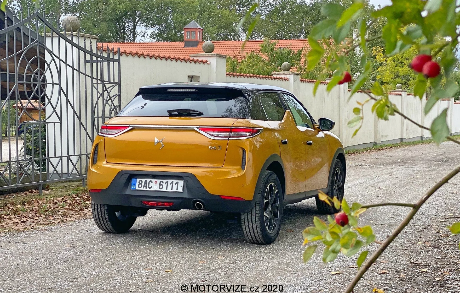 Widok z tylnego kąta DS Automobiles DS 3 Crossback 2019, uchwycony z niskiej perspektywy na podjeździe rezydencji. Samochód posiada wyraźne poziome tylne światła, centralnie umieszczone logo producenta na rzeźbionym bagażniku z wbudowanym spojlerem i trzecim światłem stop, centralnie umieszczoną tablicę rejestracyjną oraz błyszczące metaliczne wykończenie w kolorze pomarańczowym lub złotym. Solidny tylny zderzak ma zintegrowane czujniki lub reflektory, widoczny tylny wycieraczkę, antenę w kształcie płetwy rekina i wieloszprychowe aluminiowe koła. Lekko rozmazana roślinność na pierwszym planie kontrastuje z ostrzejszym punktem skupienia na samochodzie i bramie wejściowej w tle, co sugeruje miękkie rozproszone światło dzienne.