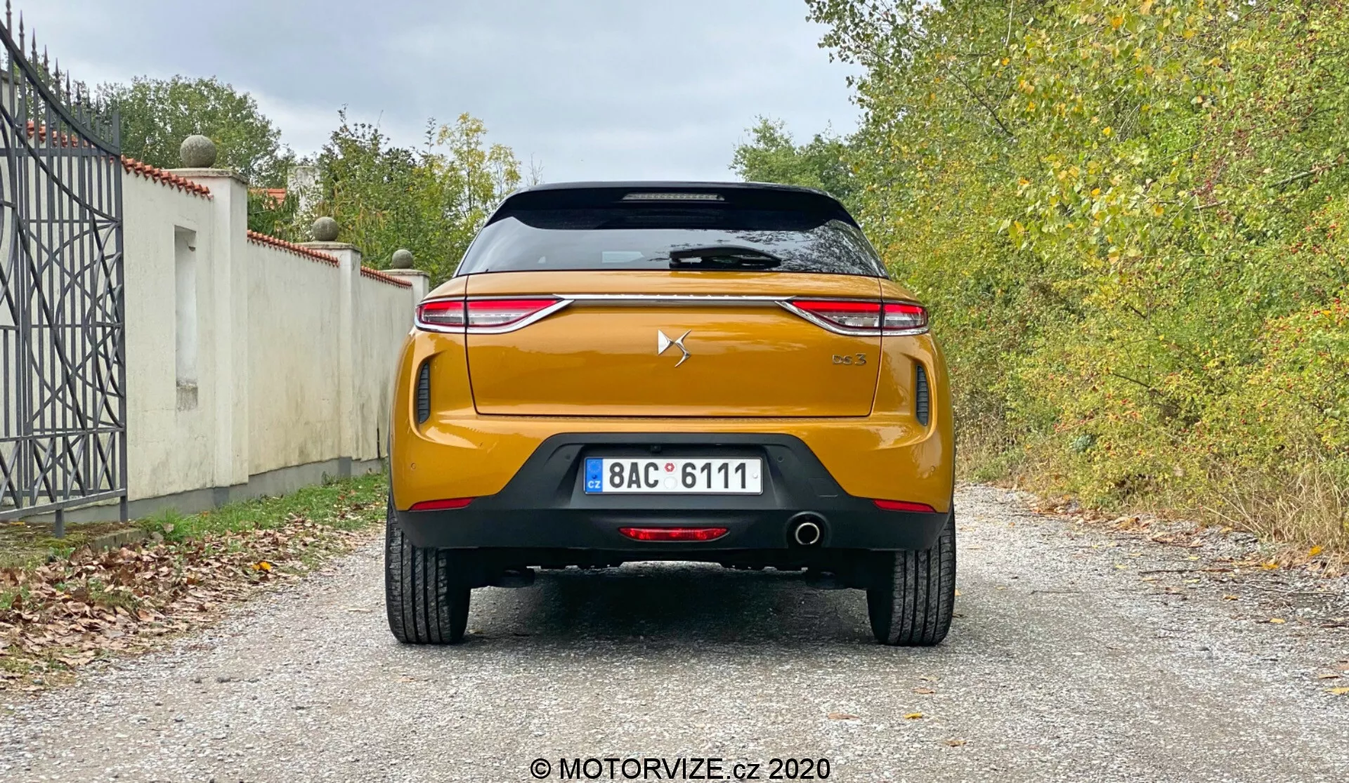 Tylny widok nowoczesnego samochodu DS-Automobiles DS-3 Crossback 2019 pokazujący jego cechy projektowe i technologiczne, w tym spoiler dachowy, tylną szybę z elementem grzewczym, wysokie światło stop, pokrywę bagażnika z logo producenta, wyraźny pasek chromowany, eleganckie tylne światła LED, solidny tylny zderzak z czujnikami parkowania, podwójny układ wydechowy, czarny element dyfuzorowy i uderzający pomarańczowy lub miedziany kolor na żwirowej powierzchni z roślinnością w tle.