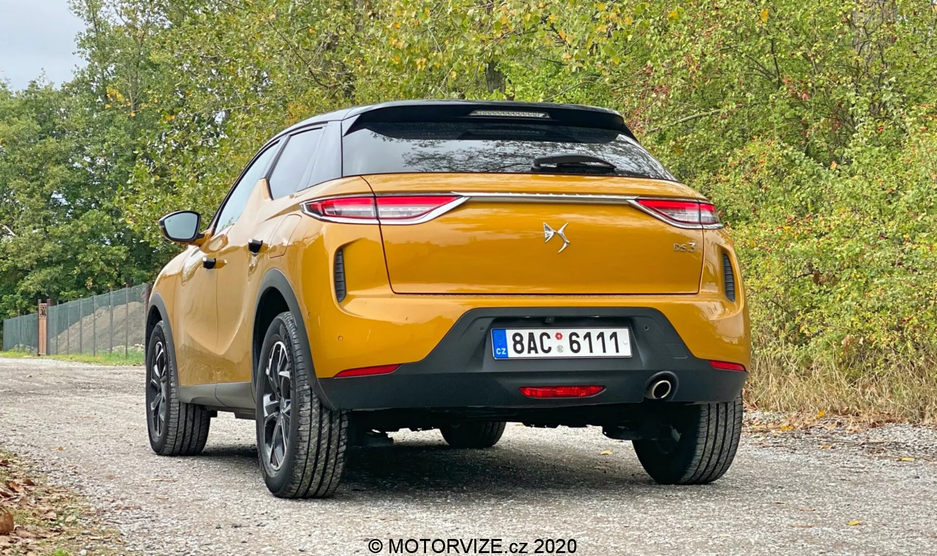 Zdjęcie przedstawia widok z tyłu współczesnego, złotego DS-3 Crossback 2019, zaparkowanego na nieutwardzonym poboczu z roślinnością po obu stronach. Samochód, uchwycony z niskiego kąta, prezentuje ostre tylne światła skierowane w stronę środka bagażnika, stylizowany emblemat chevron z sylwetką lwa na klapie bagażnika, solidny tylny zderzak z czujnikami parkowania, podwójne rury wydechowe, czarne plastikowe listwy i pięcioramienne obręcze aluminiowe. Tablica rejestracyjna jest w stylu europejskim, ze standardowymi tylnymi odblaskami i tylnym światłem przeciwmgielnym. Miękkie, rozproszone oświetlenie sugeruje pochmurny dzień, co dodaje swobodnej, wszechstronnej atmosfery, a znak wodny ‘MOTORVIZE.cz 2020’ wskazuje na źródło zdjęcia.