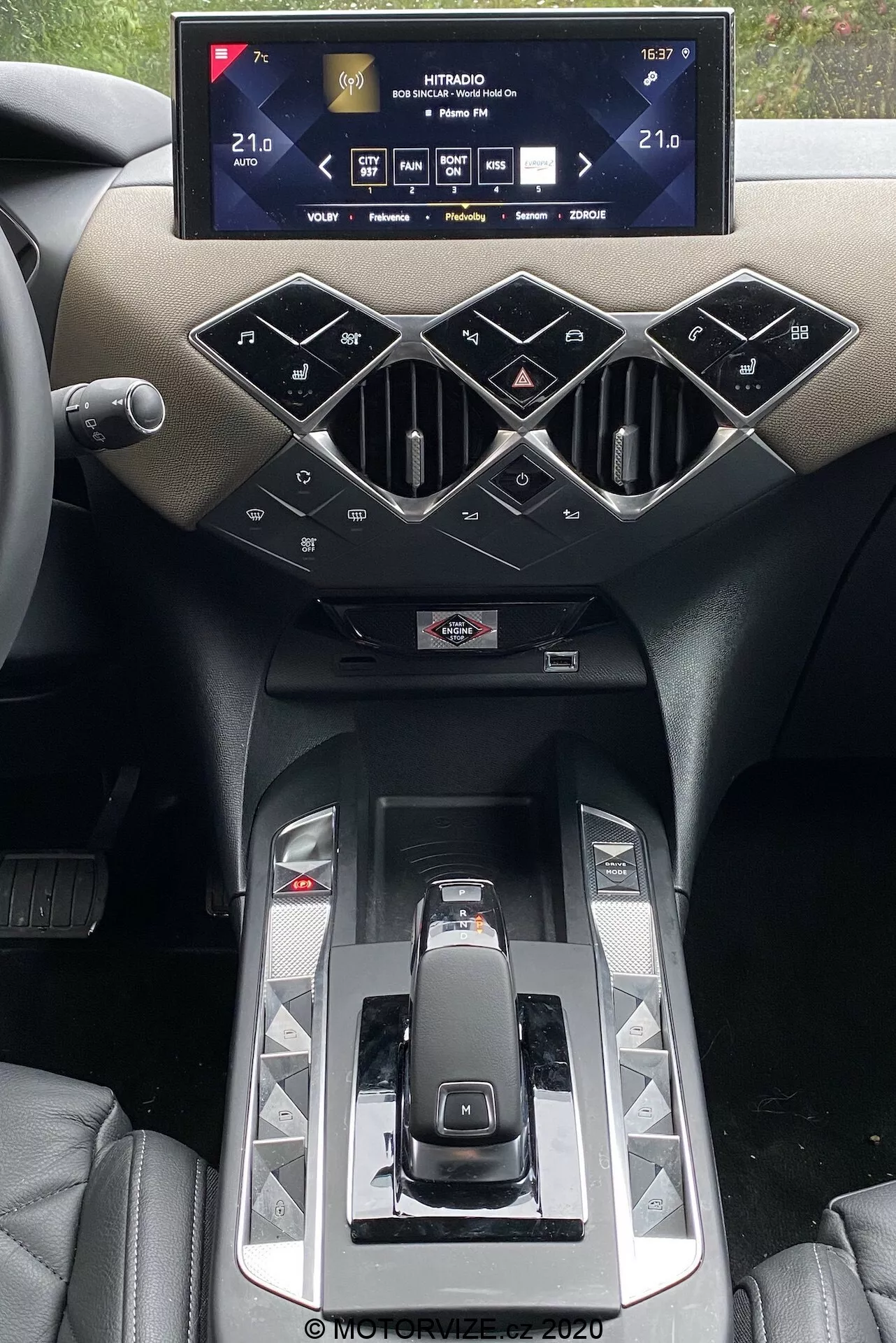 Wnętrze DS Automobiles DS 3 2019 koncentrujące się na konsoli środkowej z systemem infotainment. Szeroki, poziomo zorientowany ekran dotykowy wyświetla ustawienia pojazdu, w tym temperatury klimatyzacji ustawione na 21,0°C. Poniżej znajdują się geometrycznie stylizowane kratki wentylacyjne i fizyczne przyciski w układzie diamentowym do sterowania systemami samochodu. Nowoczesna dźwignia zmiany biegów, błyszczące panele i widoczny uchwyt na napoje podkreślone są luksusowymi materiałami i eleganckim designem.