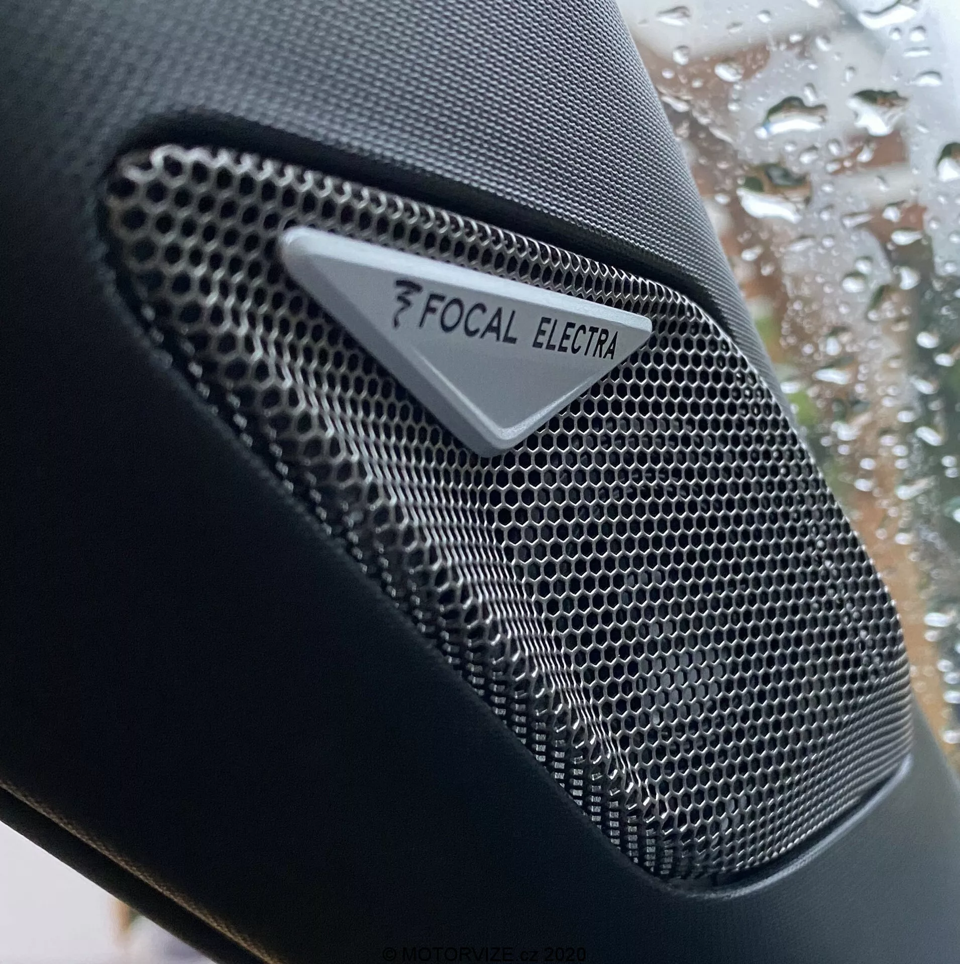 Zbliżenie głośnika marki Focal Electra w DS Automobiles DS-3 Crossback 2019, ukazujące wysokiej jakości komponent audio z metalową kratką o strukturze plastra miodu i wyraźnym logo Focal oraz marką Electra. Zdjęcie koncentruje się na złożonym projekcie głośnika, z małą głębią ostrości, która podkreśla teksturę głośnika na rozmytym tle okna samochodu z kroplami deszczu.