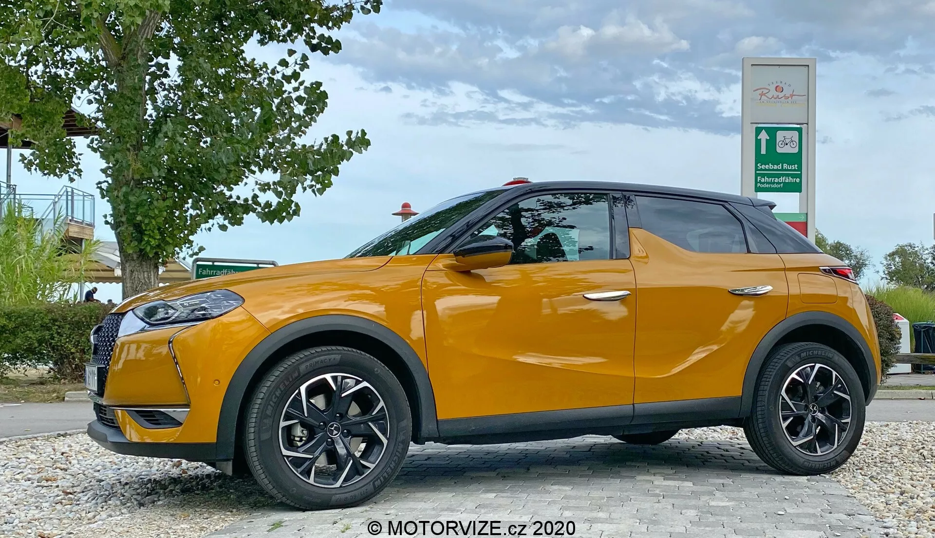 Profil boczny DS Automobiles DS 3 Crossback 2019 w jasnopomarańczowym kolorze, zaparkowanego w pobliżu przydrożnej okolicy z zielenią i znakiem stacji benzynowej. Strona kierowcy z czarnymi kołami wieloszprychowymi i lusterkami bocznymi w kolorze nadwozia jest wyraźnie widoczna z niskiego kąta na wysokości kół auta, pokazując elegancki design kompaktowego SUV-a z rosnącą linią dolną okien i przyciemnianymi szybami.