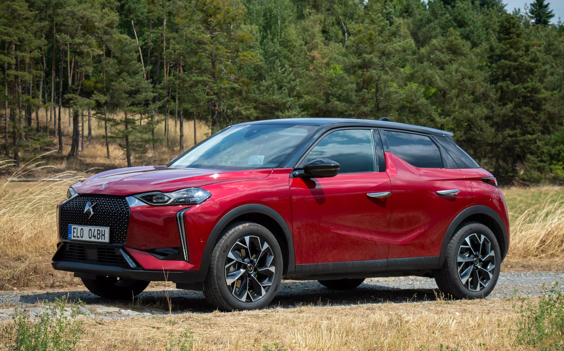 Widok z boku przedni DS Automobiles DS 3 E-TENSE 2023, czerwony crossover SUV, felgi aluminiowe wieloramienne, reflektory LED, na ścieżce żwirowej z tłem leśnym