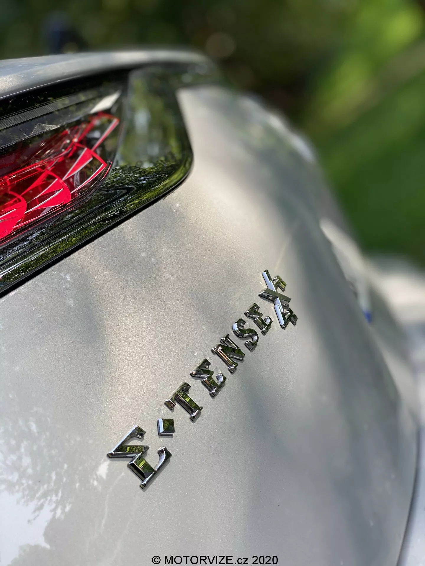Zbliżenie tylnej części modelu DS Automobiles DS 7 Crossback 2018, skupiające się na emblemacie 'E-TENSE 4X4', który wskazuje na napęd na cztery koła i prawdopodobnie hybrydowy lub elektryczny układ napędowy. Płytka głębia ostrości rozmywa tło, jednocześnie uwydatniając emblemat, z widocznym powyżej czerwonym, krystalicznym światłem tylnym. Obraz przedstawia metaliczne wykończenie nadwozia i chromowane litery w naturalnym świetle słonecznym, podkreślając zaawansowaną technologię i luksusowy design pojazdu.