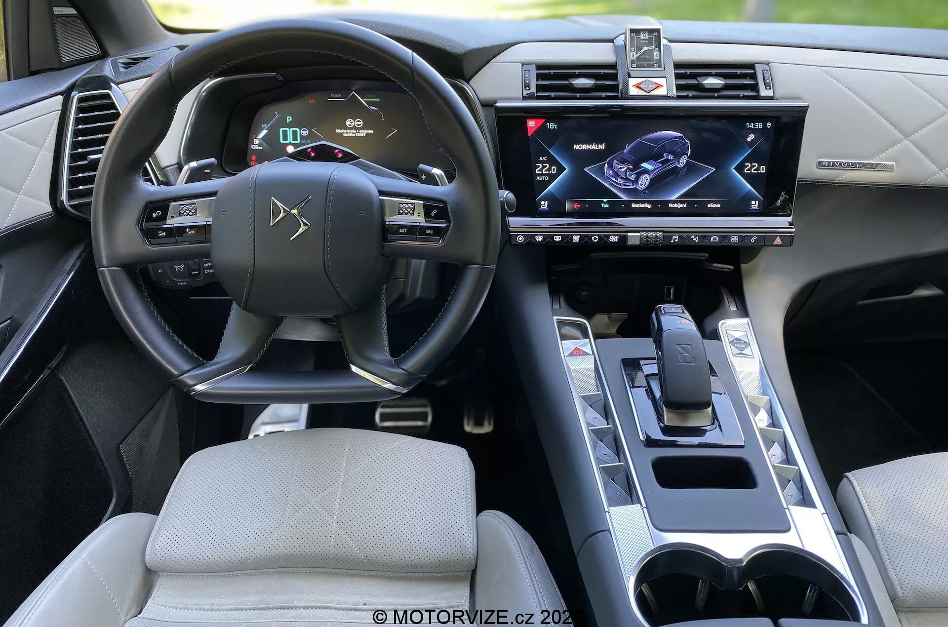 Wnętrze DS Automobiles DS 7 Crossback 2018, widok z tylnego siedzenia skupiający się na stronie kierowcy. Funkcje obejmują kierownicę z wbudowanymi sterownikami, cyfrowy zestaw wskaźników, centralnie zamontowany dotykowy system informacyjno-rozrywkowy, nowoczesny drążek zmiany biegów, uchwyty na kubki i wyrafinowany design wnętrza z zaawansowanymi udogodnieniami technologicznymi.