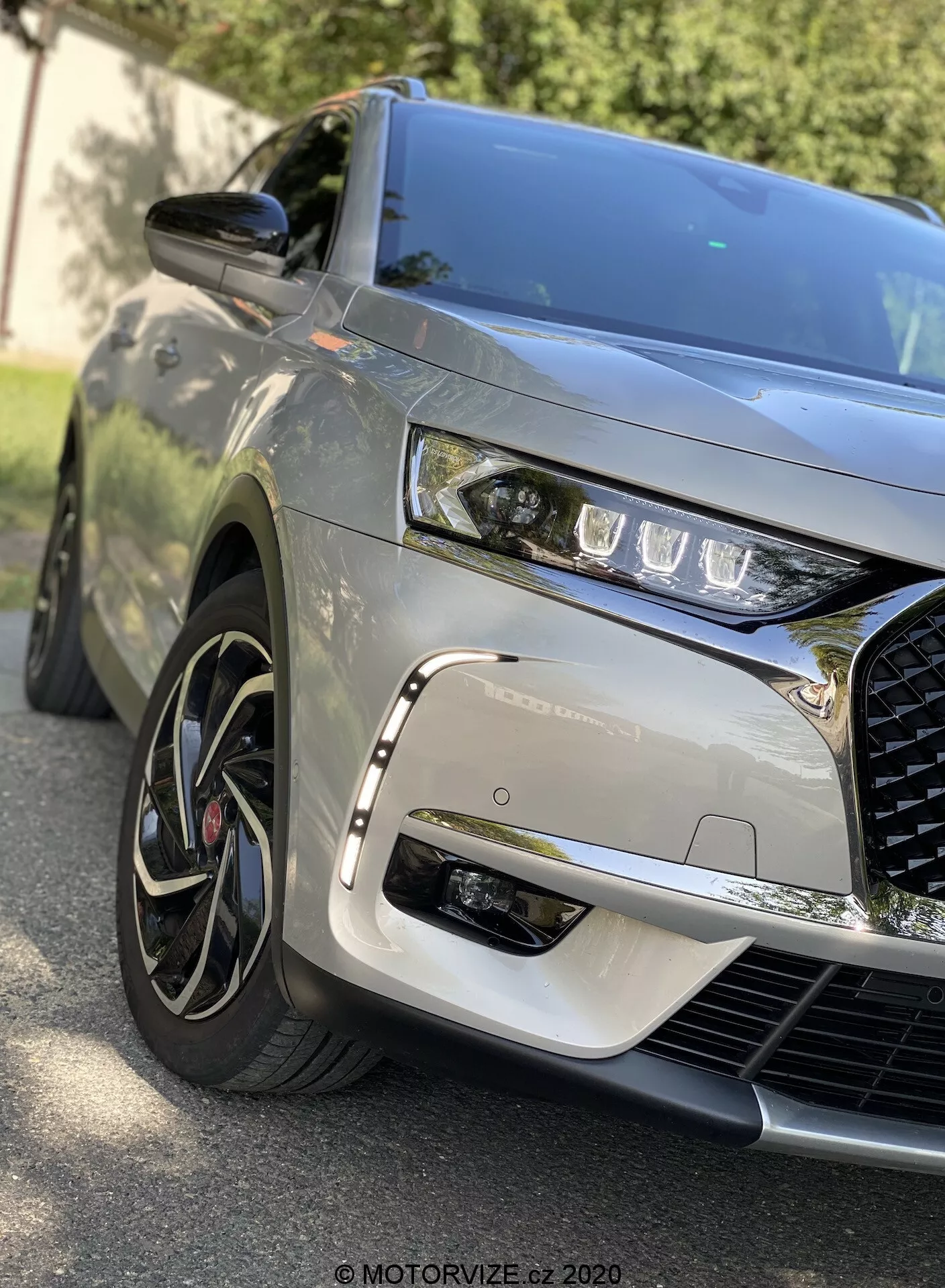 Zbliżenie przodu z przodu DS Automobiles DS 7 Crossback 2018, ukazujące lewą przednią ćwiartkę, ukazujące eleganckie reflektory LED, dominującą złożoną kratkę, wyrzeźbioną maskę, wieloszprychowe alufelgi z oponami o niskim profilu, metaliczne wykończenie lakieru, boczne lusterko w kolorze nadwozia i dynamiczne odbicia wskazujące na otoczenie zewnętrzne.