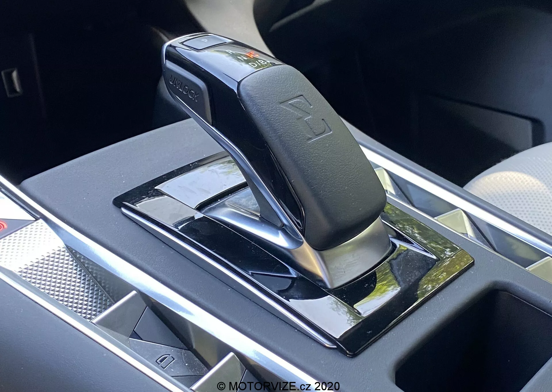 Zbliżenie dźwigni automatycznej skrzyni biegów DS Automobiles DS-7 2018 (DS-7 Crossback), pokazujące lśniącą czarną dźwignię ze srebrnymi akcentami, prawdopodobnie w pozycji park lub neutralnej, zamontowaną na czarnym fortepianowym panelu z refleksyjnymi powierzchniami. Funkcje obejmują wytłoczone symbole pozycji biegów, przycisk bezpieczeństwa na dźwigni oraz pobliski schowek z potencjalną bezprzewodową ładowarką. Obszar wokół ma chromowane lub srebrne wykończenia, wskazujące na nowoczesne i luksusowe wnętrze pojazdu.