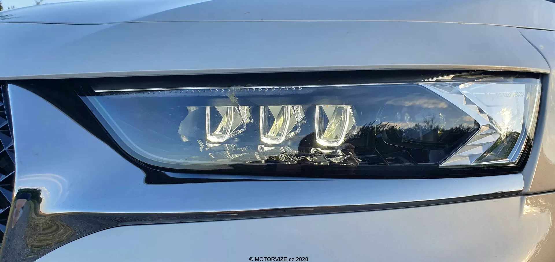 Zbliżenie lewej lampy samochodu DS Automobiles DS-7 Crossback pod lekkim kątem, ukazujące nowoczesny projekt LED z wieloma soczewkami projektorowymi oraz charakterystyczne LED-owe światła dzienne obudowane w przejrzysty, wysokiej jakości plastik. Lampa przedstawia zaawansowaną technologię oświetlenia pojazdów, zdolną do zapewnienia zarówno świateł mijania, jak i drogowych, z potencjalnymi funkcjami adaptacyjnymi poprawiającymi widoczność. Widać również subtelne odbicie na błyszczącej powierzchni i odcień jasnoszarego do średnioszarego koloru samochodu.