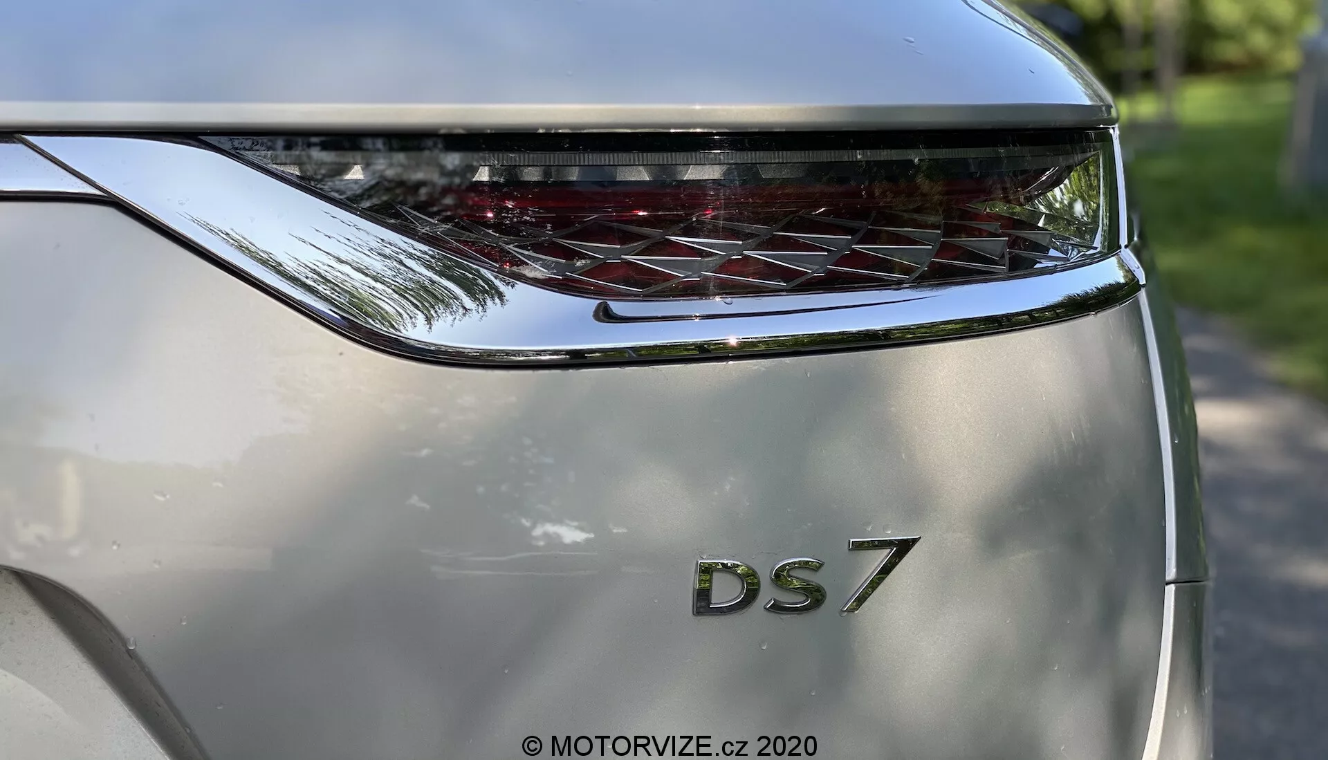Zbliżony widok tylnej sekcji DS Automobiles DS 7 Crossback 2018 uchwycony z niskiego kąta. Głównym punktem jest tylna klapa, na której widnieją pogrubione litery 'DS' nad oznaczeniem modelu '7'. Nad nazwą modelu znajduje się poziomo rozciągnięty, zakrzywiony tylne światło z charakterystycznymi elementami LED. Kolor nadwozia to metaliczny srebrny, a rzeźbiony design sugeruje nowoczesny i luksusowy wygląd. Fotografia podkreśla markę i design tylnego światła, z subtelnym odbiciem jasnego otoczenia zewnętrznego na powierzchni samochodu.