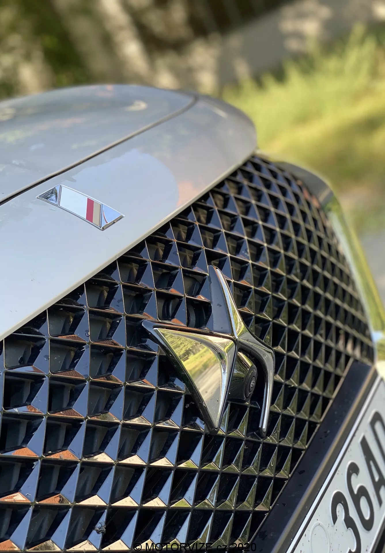 Zbliżenie na przednią lewą sekcję DS Automobiles DS 7 Crossback 2018. Obraz koncentruje się na luksusowej, skomplikowanej kratce przedniej z refleksyjnymi powierzchniami i wzorem diamentowym, z wyeksponowanym stylizowanym logo na środku. Nad kratką, maska eksponuje mniejszy emblemat znaczący model wyższej klasy. Tło jest rozmyte, podkreślając design kratki i elementy marki.