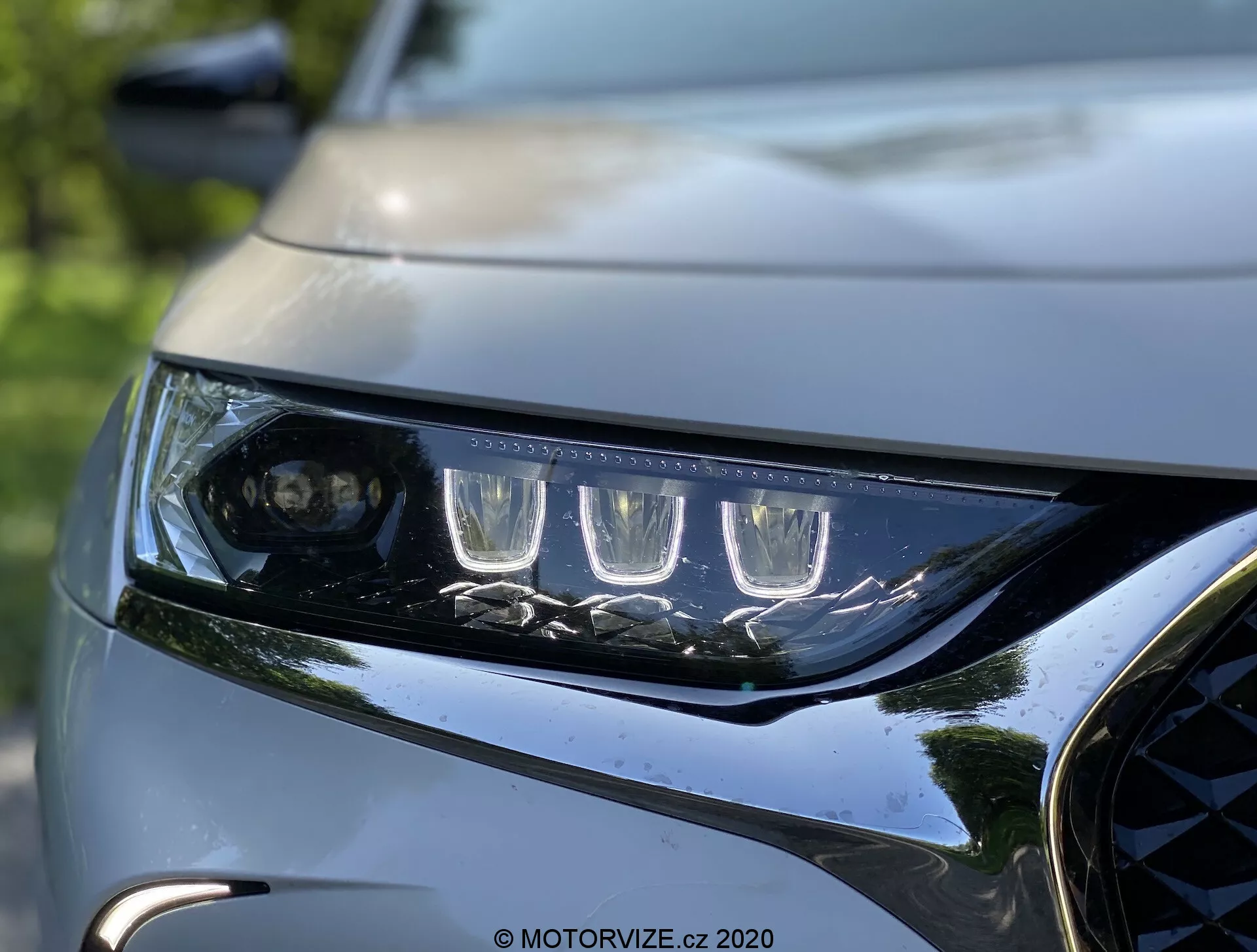Zbliżony widok lewego reflektora i przedniego grilla DS Automobiles DS 7 Crossback 2018. Reflektor ma charakterystyczne diody LED do jazdy dziennej i soczewkę projektora dla głównego strumienia światła, otoczony przez kilka elementów LED. Elegancka i kątowa obudowa reflektora pokazuje nowoczesną aerodynamikę. Część ciemnego, błyszczącego grilla z metalicznymi akcentami oraz część rzeźbionej maski podkreślają dynamiczny, premium design pojazdu. Tło jest rozmyte, podkreślając ostrość i skomplikowane detale oświetlenia i fasady przedniej części samochodu.