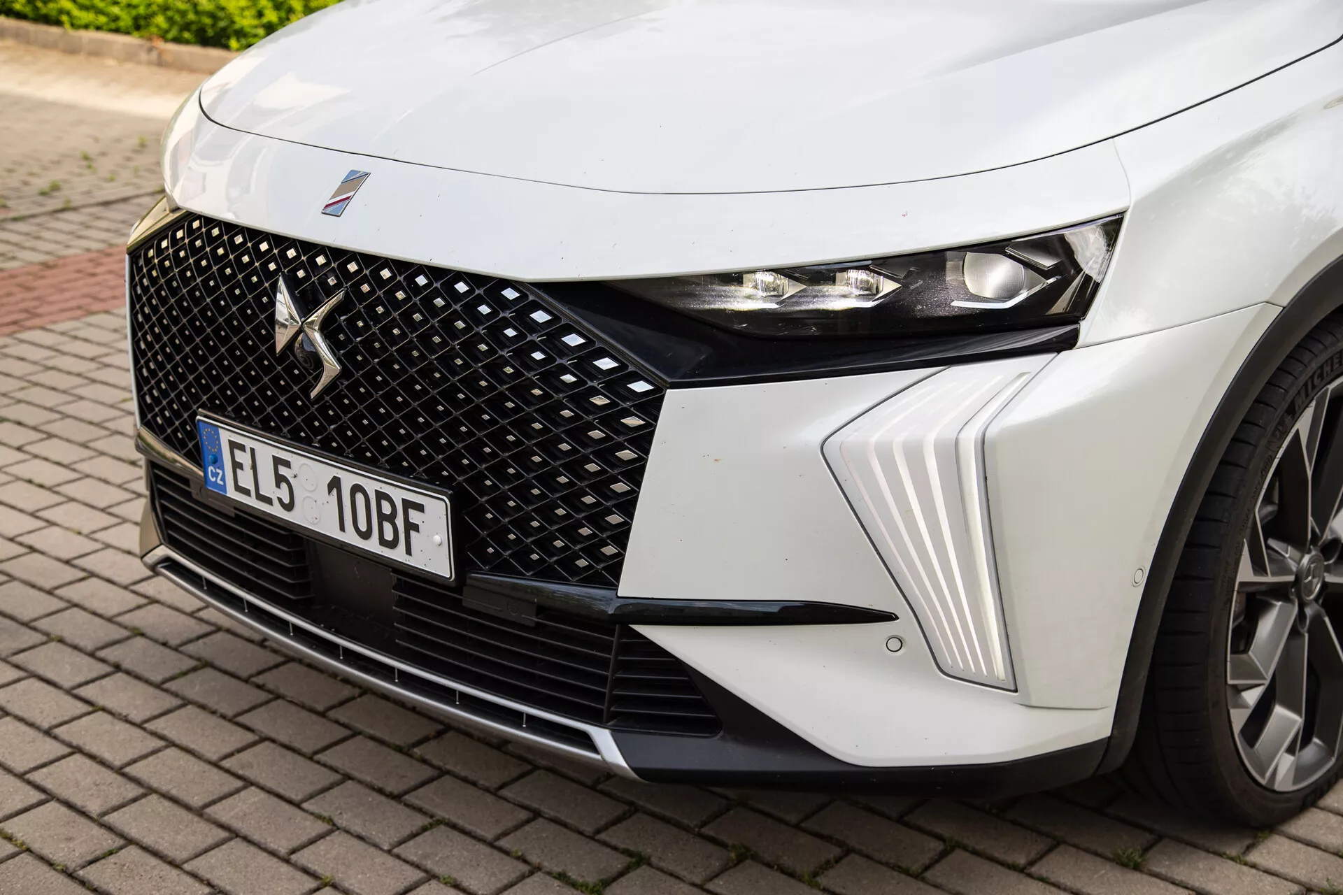 Zbliżenie na przedni lewy kwadrant DS Automobiles DS-7 2022 z szczegółowym skupieniem na reflektory LED, sześciokątny design grilla z centralną odznaką marki oraz dynamiczne linie maski wraz ze wglądem w koło ze stopu
