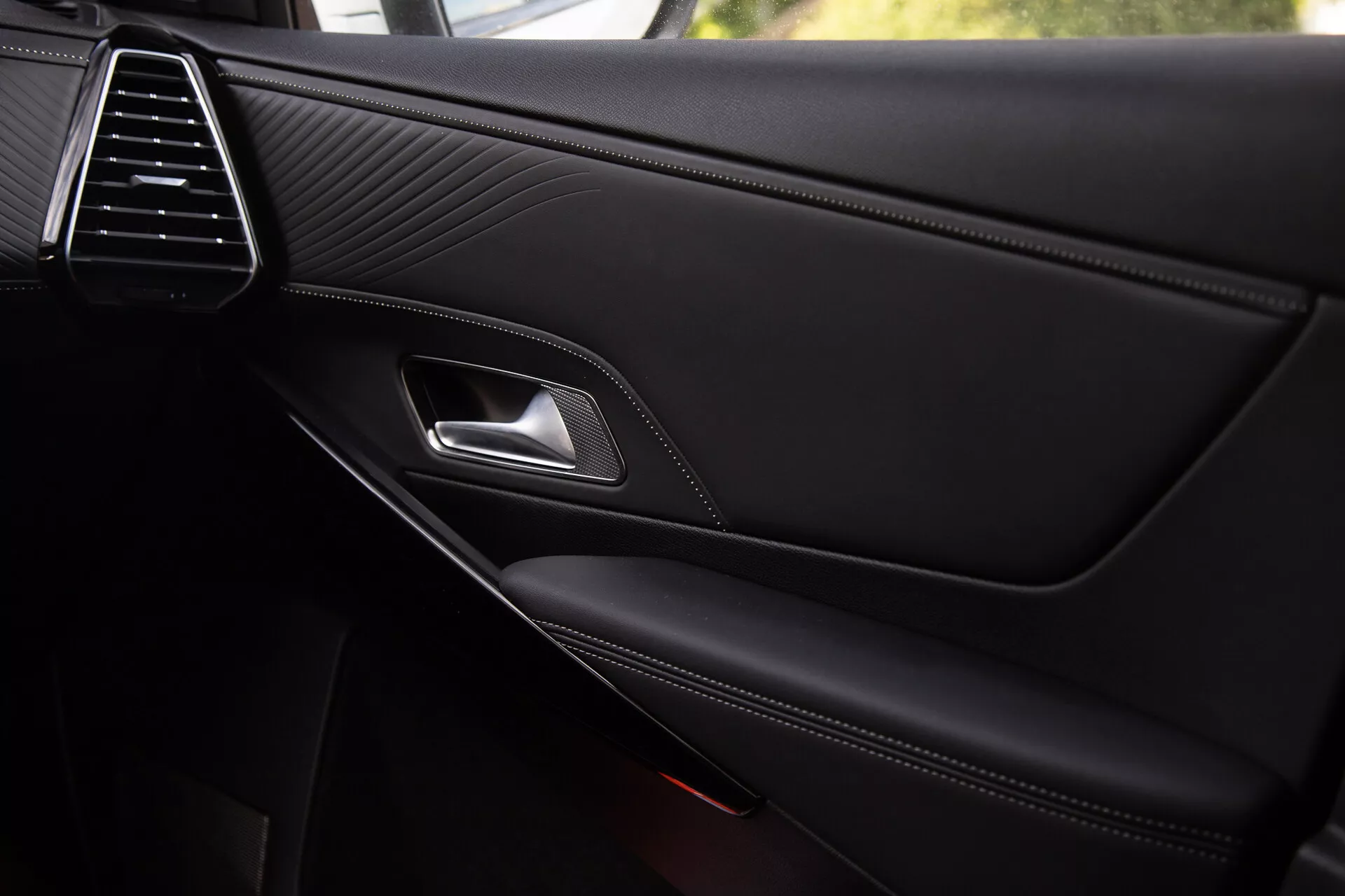 Zbliżenie na panel wewnętrzny drzwi DS Automobiles DS-7 2022 ukazujące elegancką kratkę wentylacyjną, skórzaną fakturę ze szwami, chromowaną klamkę, ergonomiczną dźwignię zatrzasku, podłokietnik z kontrastowymi szwami i wbudowaną kieszeń na drobiazgi z częściowo widoczną kratką głośnika, odzwierciedlające jakość premium i szczegóły konstrukcyjne.
