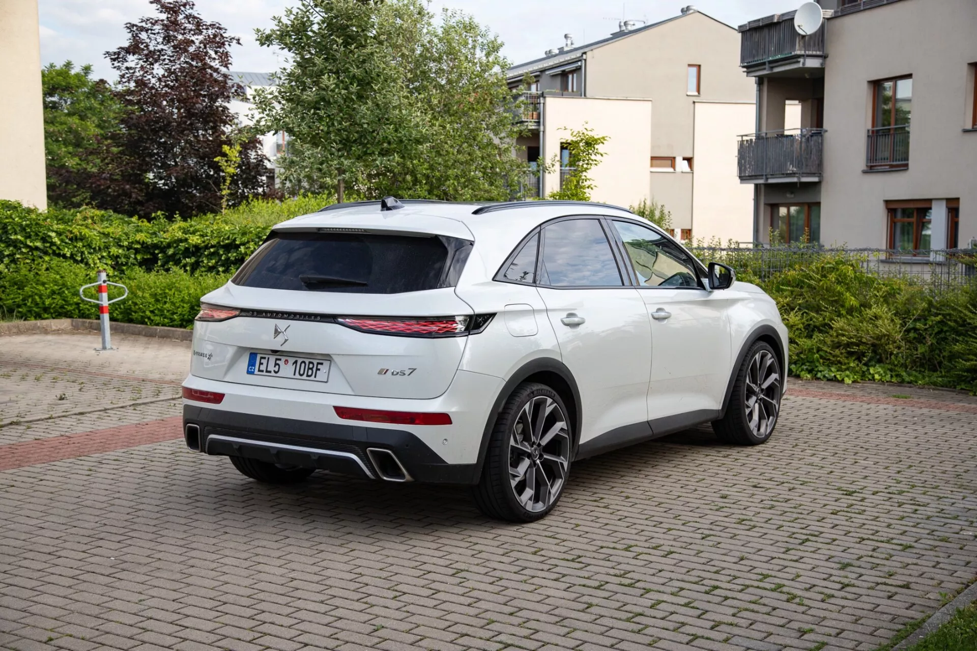 Biały SUV DS Automobiles DS-7 z 2022 roku zaparkowany na obszarze z ceglaną nawierzchnią, pokazujący tylne światła LED, podwójny układ wydechowy i sportowe koła ze stopów. Zdjęcie uchwyciło pojazd o eleganckiej, coupé-podobnej konstrukcji SUV z perspektywy trzech czwartych od tyłu, podkreślając możliwy panoramiczny dach oraz lusterka boczne z zaawansowaną technologią.