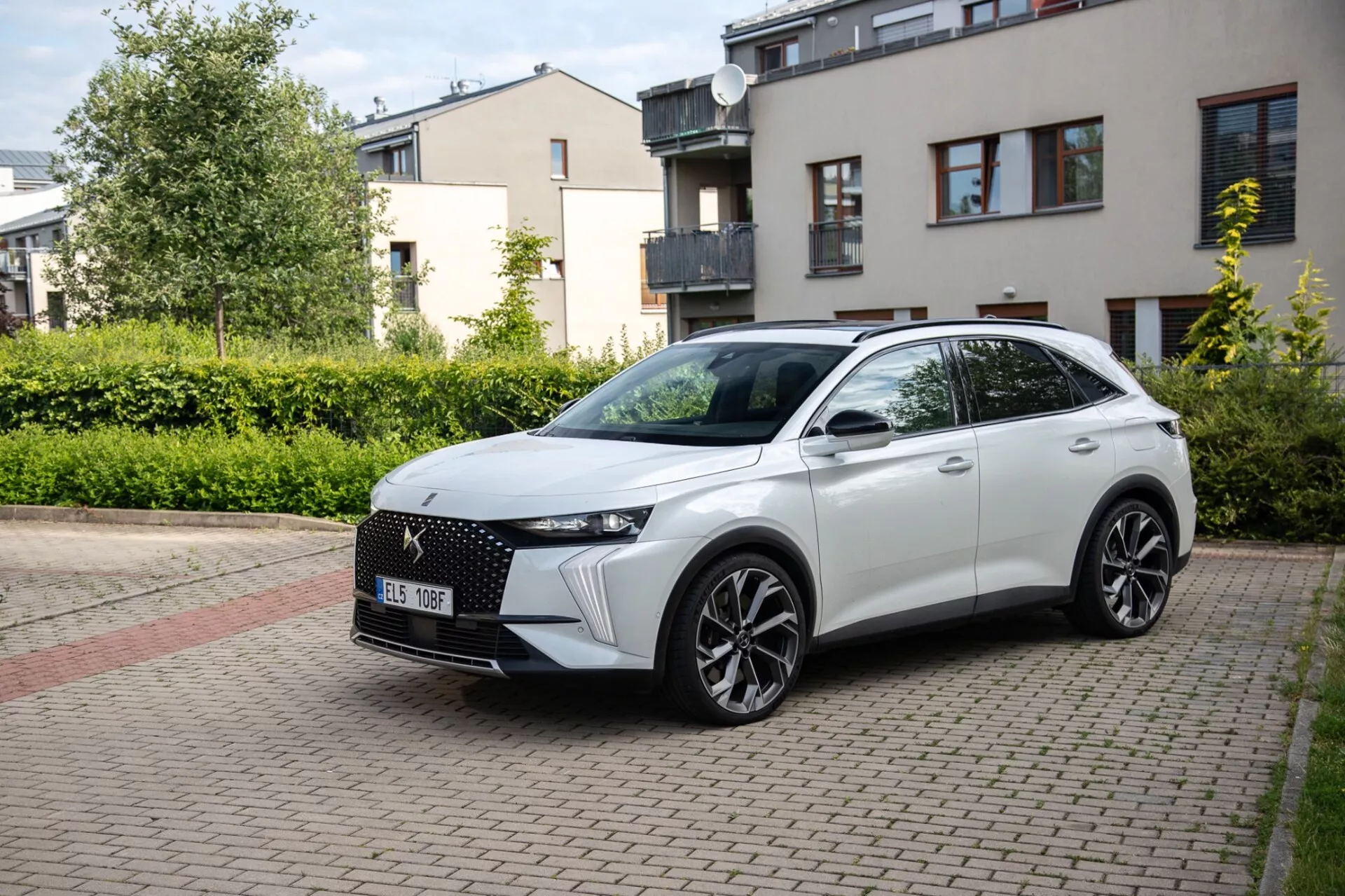Trzy czwarte widok z przodu crossovera DS Automobiles DS-7 2022 zaparkowanego na brukowanej kamieniem ulicy mieszkalnej, prezentującego jego nowoczesny design łącznie z charakterystycznym przednim grillem, eleganckimi reflektorami LED, rzeźbioną maską, dużymi kołami ze stopu i sportowym profilem bocznym z wbudowanymi kierunkowskazami na lusterkach bocznych montowanych na drzwiach pod naturalnym światłem dziennym.