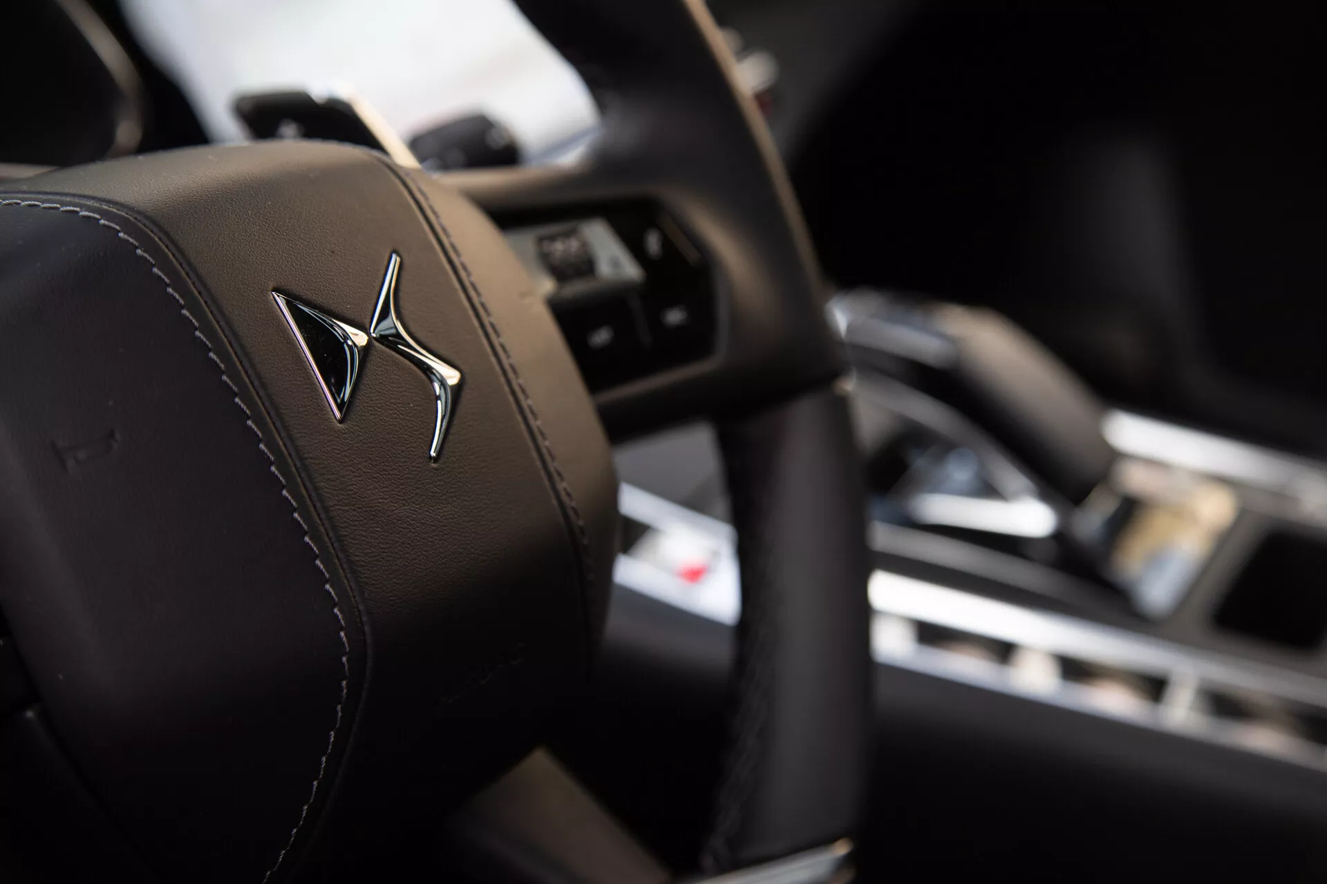 Zbliżenie na zagłówek siedzenia kierowcy z logo w tłoczeniu, ze ś Shallow depth of field highlighting the headrest in sharp focus against a blurred interior background showcasing a steering wheel with control buttons and a dashboard with a central display screen.allow głębią pola podkreślająca zagłówek w ostrej ostrości w kontraście do rozmazanego tła wnętrza prezentującego kierownicę z przyciskami sterującymi i deski rozdzielczej z centralnym ekranem.