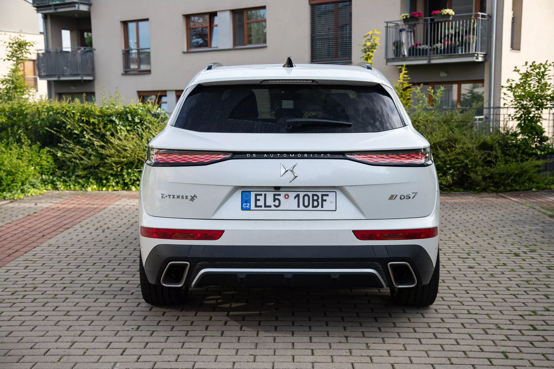 Tylny widok DS Automobiles DS 7 2022 zaparkowanego na nawierzchni z cegieł z budynkami i zielenią w tle, prezentujący pasek świetlny LED, tylne światła, podwójne wyjścia wydechowe i tablicę rejestracyjną UE
