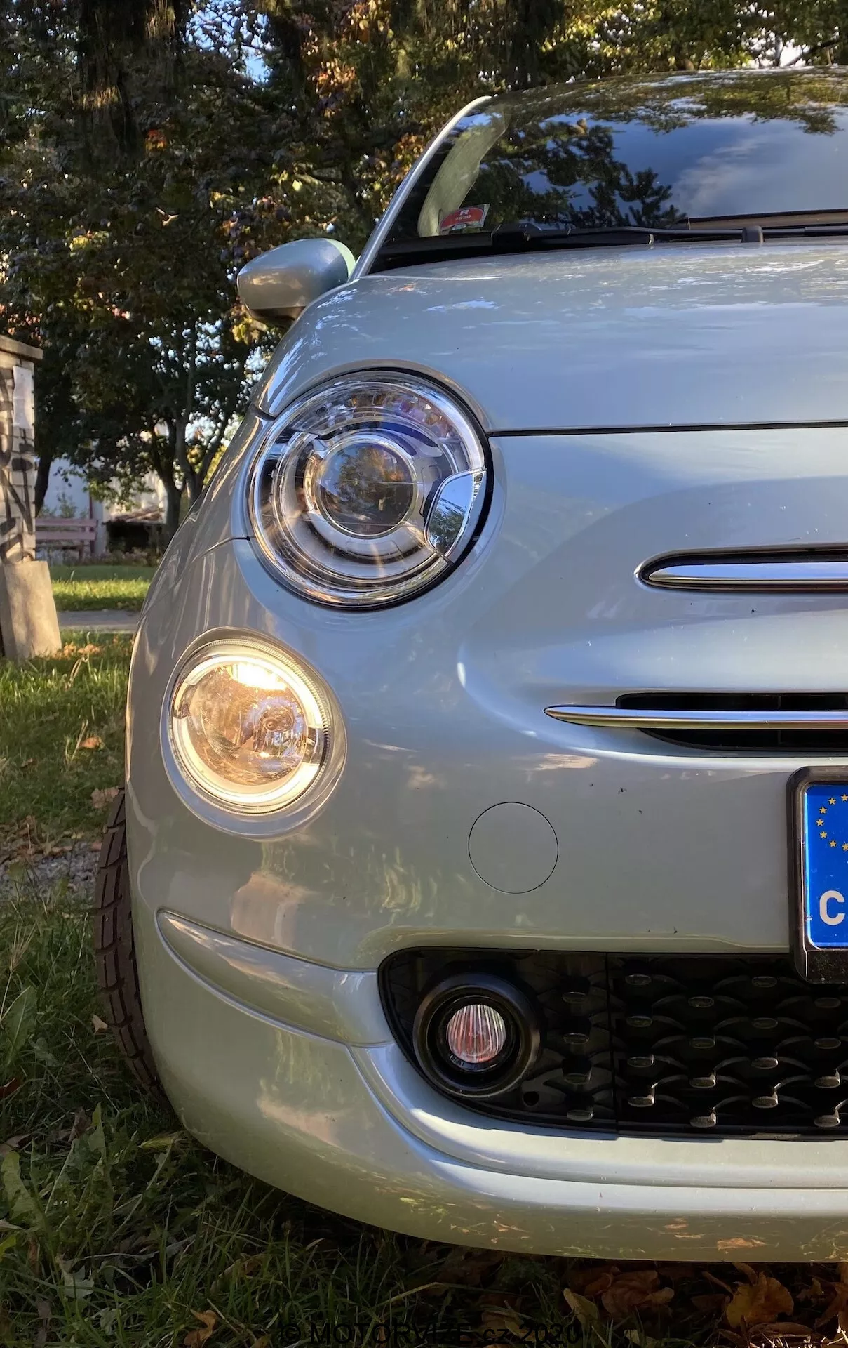 Zbliżenie przedniego lewego narożnika Fiata 500 z 2015 roku, uchwyconego pod niskim kątem, uwidaczniającego maskę, przedni zderzak, nadkole i okrągły reflektor z projektorem otoczonym pierścieniem DRL. Zestaw zawiera pomarańczowy kierunkowskaz oraz światło przeciwmgielne poniżej. Chromowane akcenty zdobią obręcz reflektora, obudowę światła przeciwmgielnego i dwie poziome listwy na grillu. Grill ma czarny wzór w kształcie plastra miodu. Zaokrąglony przód i tablica rejestracyjna w stylu europejskim wskazują na retro-nowoczesny design auta, zaparkowanego na trawie z liśćmi jesiennymi.