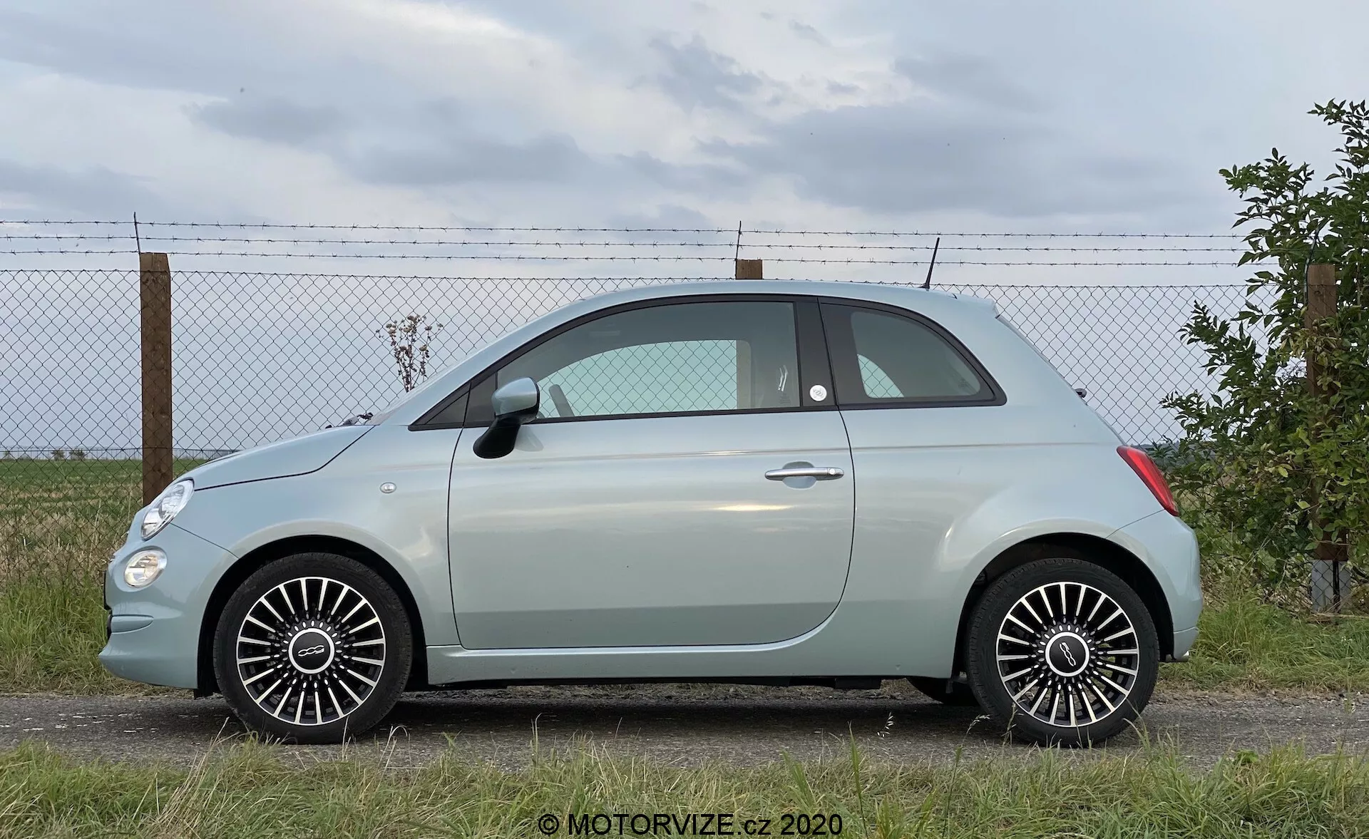 Nowoczesny subkompaktowy hatchback z bocznego profilu, pokazujący całą długość pojazdu. Zaparkowany na równym podłożu z ogrodzeniem i horyzontem z chmurami w tle. Cechy: boczne drzwi, okna, lusterka, konfiguracja dwudrzwiowa, małe tylne okienko, zaokrąglony dach, pastelowy kolor nadwozia z kontrastującym dachem, wieloramienne felgi aluminiowe, opony w dobrym stanie, szczegółowe linie projektowe i naturalne oświetlenie dzienne.