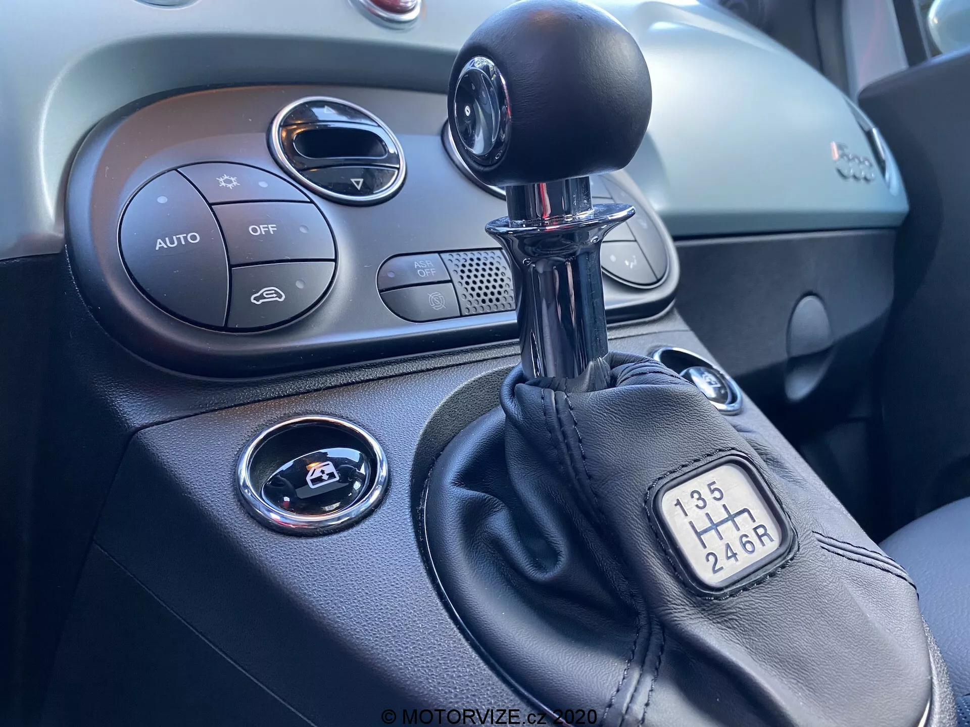 Zbliżenie konsoli centralnej wewnątrz Fiata 500 z 2015 roku, koncentrujące się na prawej stronie. Obraz pokazuje manualną dźwignię zmiany biegów z konfiguracją 6-biegową, część konsoli centralnej z kontrolami klimatyzacji oznaczonymi 'AUTO', przycisk 'OFF', przycisk start/stop do zapłonu, okrągły otwór wentylacyjny i teksturowaną deskę rozdzielczą.