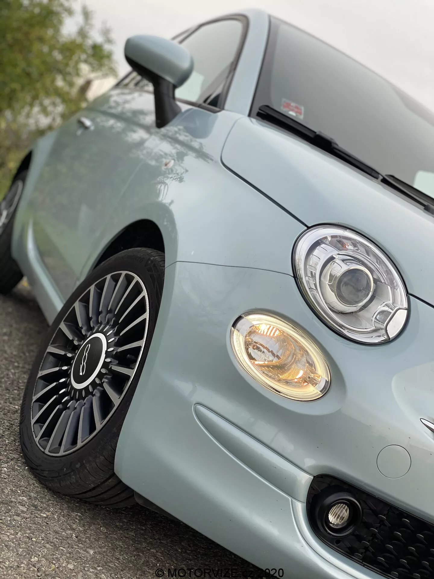 Zbliżenie na przedni lewy kwartał jasnoniebieskiego Fiata 500 z 2015 roku, ukazujące nowoczesny reflektor projektorowy z pierścieniem LED lub halo, podświetlony reflektor przeciwmgielny, sportowy grill z siatką w kształcie plastra miodu, przednią część maski oraz wieloszprychowe koło aluminiowe z głębokim bieżnikiem, sugerujące dobrze utrzymany pojazd i zaawansowany design motoryzacyjny.