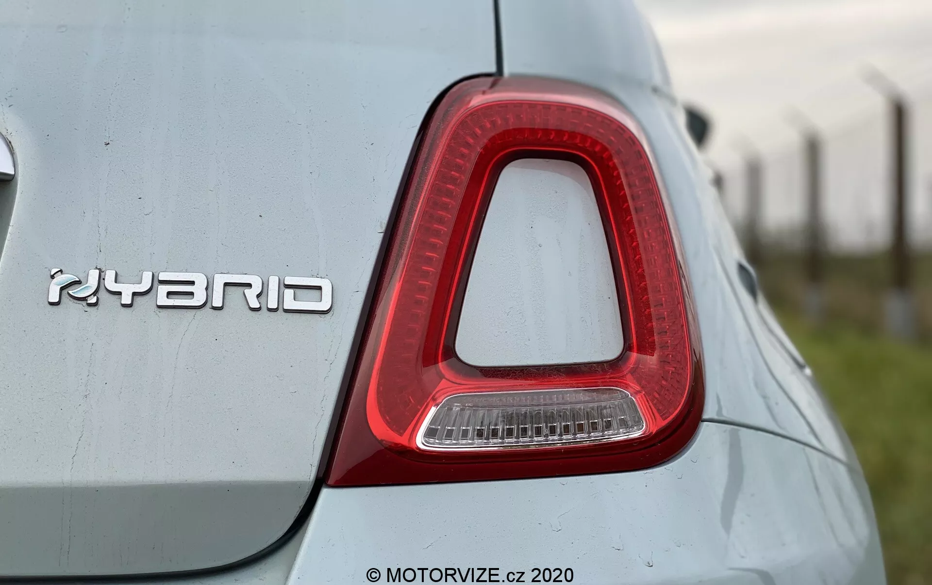 Zbliżenie na prawe tylne światło i odznakę 'HYBRID' na Fiacie 500 z 2015 roku. Tylne światło ma kształt odwróconej litery L, jest głównie czerwone z przezroczystą sekcją dla światła cofania, i jest zintegrowane z karoserią samochodu. Metaliczna odznaka 'HYBRID', oznaczająca technologię hybrydową, jest wyraźnie umieszczona. Jasny kolor nadwozia ma odbijającą jakość z niektórymi plamami kurzu lub wody, co sugeruje zewnętrzne otoczenie. Zdjęcie zrobione jest z lekko niskiego kąta, podkreślając design tylnego światła i technologię hybrydową.