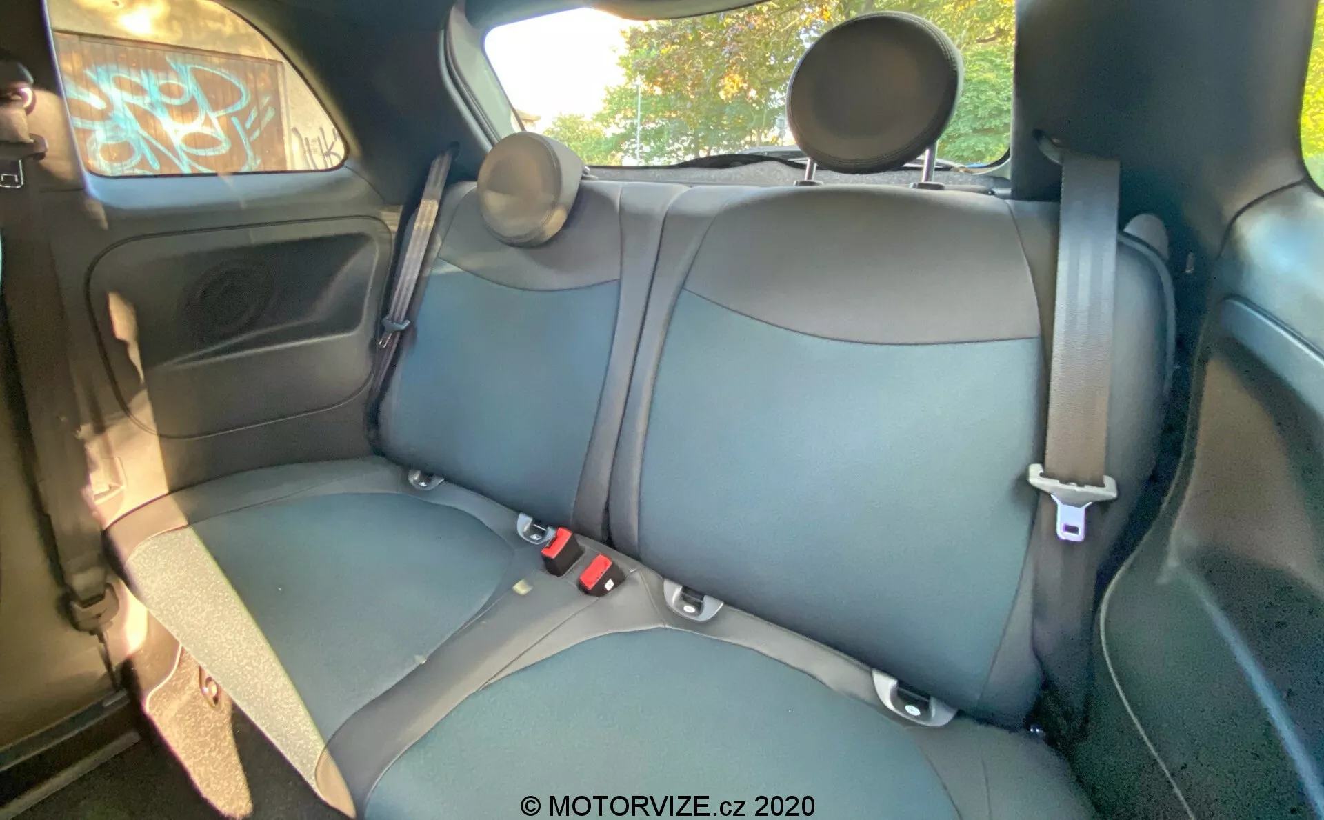 Wnętrze Fiata 500 z 2015 roku, pokazujące tylne siedzenia pasażerów z tapicerką skórzaną lub z materiału skóropodobnego w ciemnym kolorze, widoczne szwy, trzypunktowe pasy bezpieczeństwa, regulowane zagłówki, panel drzwiowy z elektrycznym sterowaniem okna, schowek, kratkę głośnika, częściowy widok sufitu i uchwyt górny. Kąt kamery jest lekko podniesiony od przodu do tyłu, zapewniając szczegółowy widok na tylną kanapę, tylne drzwi, część sufitu i okna.