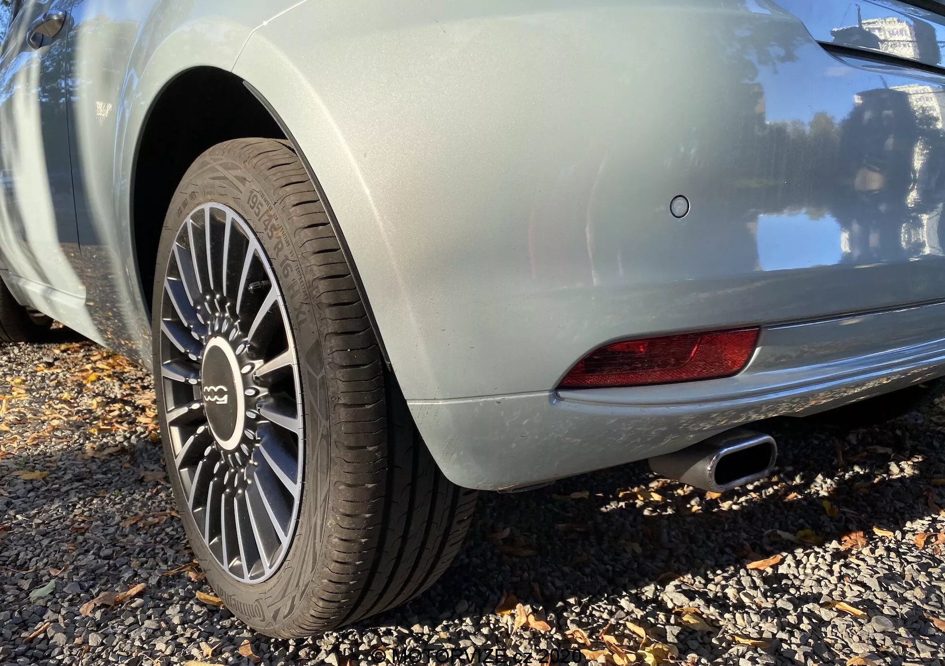 Zbliżenie tylnej części Fiat 500 2015 w jasnym kolorze, zrobione pod niskim kątem. Skupiono się na wieloramiennym kole aluminiowym i oponie z widocznym bieżnikiem, co wskazuje na sportowy model. Tylny błotnik ma elegancką krzywiznę, a metaliczna powłoka nadwozia odbija stłumiony połysk. Widoczna jest sekcja tylnego światła LED i chromowana końcówka wydechu. Czujnik parkowania i tylne światło przeciwmgielne wskazują na zaawansowane funkcje bezpieczeństwa.