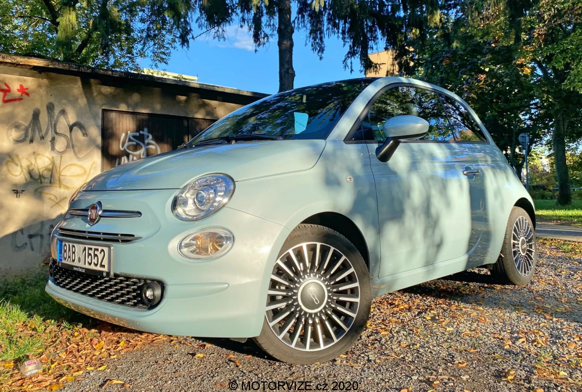 Zdjęcie pod kątem niskim przedstawiające Fiat 500 2015 z przodu bocznego, podkreślające stronę kierowcy. Obraz podkreśla grill, przedni zderzak, reflektory, światła przeciwmgielne i szczegółowe felgi aluminiowe. Samochód jest zaparkowany na powierzchni pokrytej jesiennymi liśćmi, z trawiastym terenem i graffiti w tle, oświetlony naturalnym światłem dnia, sugerując poranne lub późne popołudniowe oświetlenie.