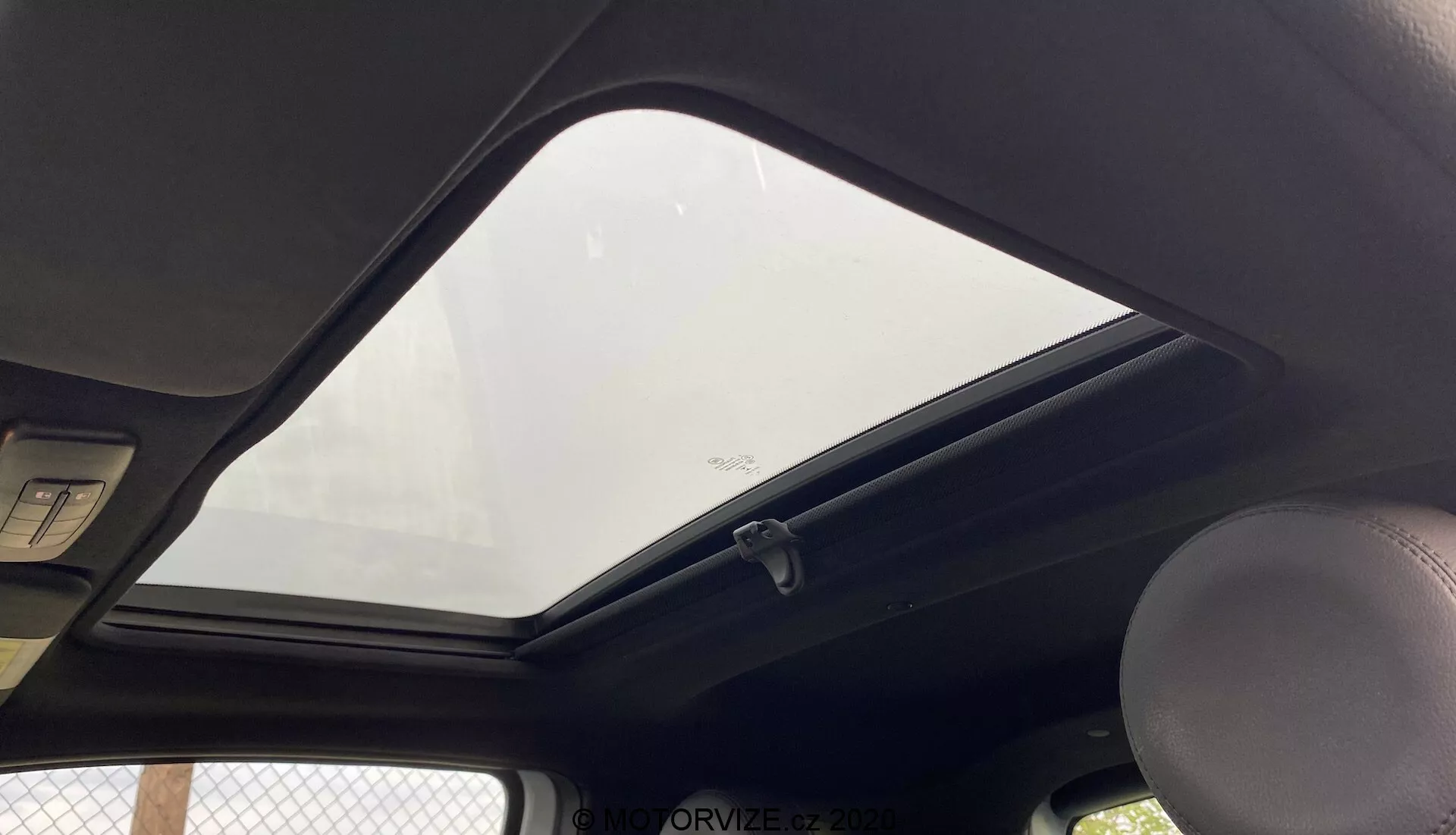 Wnętrze Fiata 500 z 2015 roku z widokiem na górną część dachu z perspektywy pasażera lub kierowcy. Zamknięty, wyeksponowany dach panoramiczny wpuszcza naturalne światło do kabiny. Ciemna wyściółka dachu kontrastuje z zewnętrzem. Widoczny uchwyt po lewej stronie, standardowe oświetlenie sufitowe powyżej oraz krawędzie dwóch osłon przeciwsłonecznych u góry. Wieszak na ubrania znajduje się w pobliżu krawędzi szyby dachu panoramicznego, na dole widoczna jest etykieta bezpieczeństwa. Zdjęcie wykonane z niskiego kąta, blisko ukazujące cechy wewnętrznego dachu.