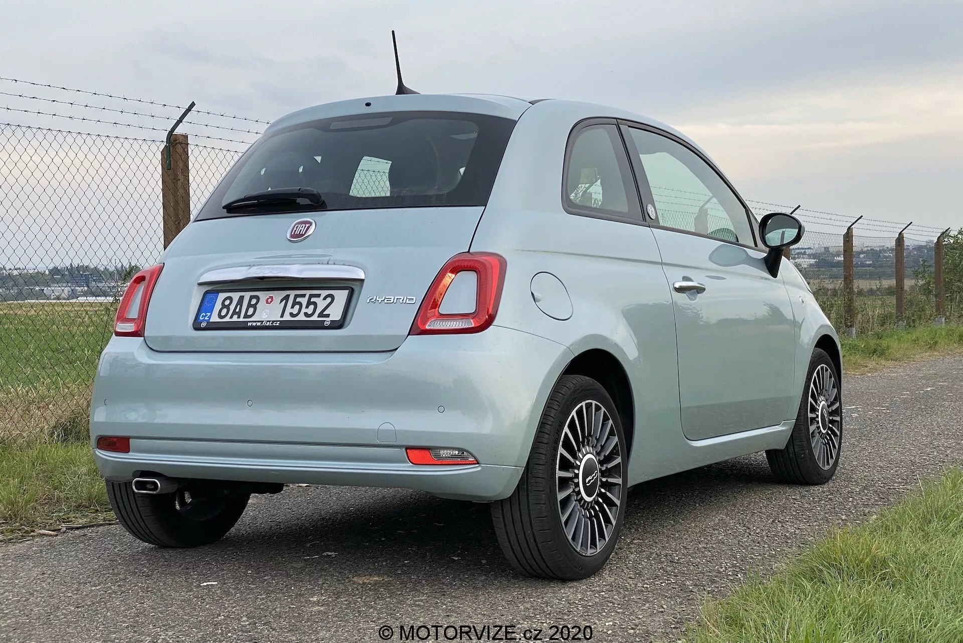 Widok zza samochodu Fiat 500 2015 w kolorze jasnoniebieskim z ujęcia pod kątem trzy czwarte, pokazujący zaokrąglone prostokątne tylne światła, tylne wycieraczki, chromowaną plakietkę, czujniki parkowania na zderzaku, rurę wydechową po lewej stronie i aluminiowe felgi. Samochód jest zaparkowany lewym bokiem do aparatu, na tle rozmytego ogrodzenia i zieleni.