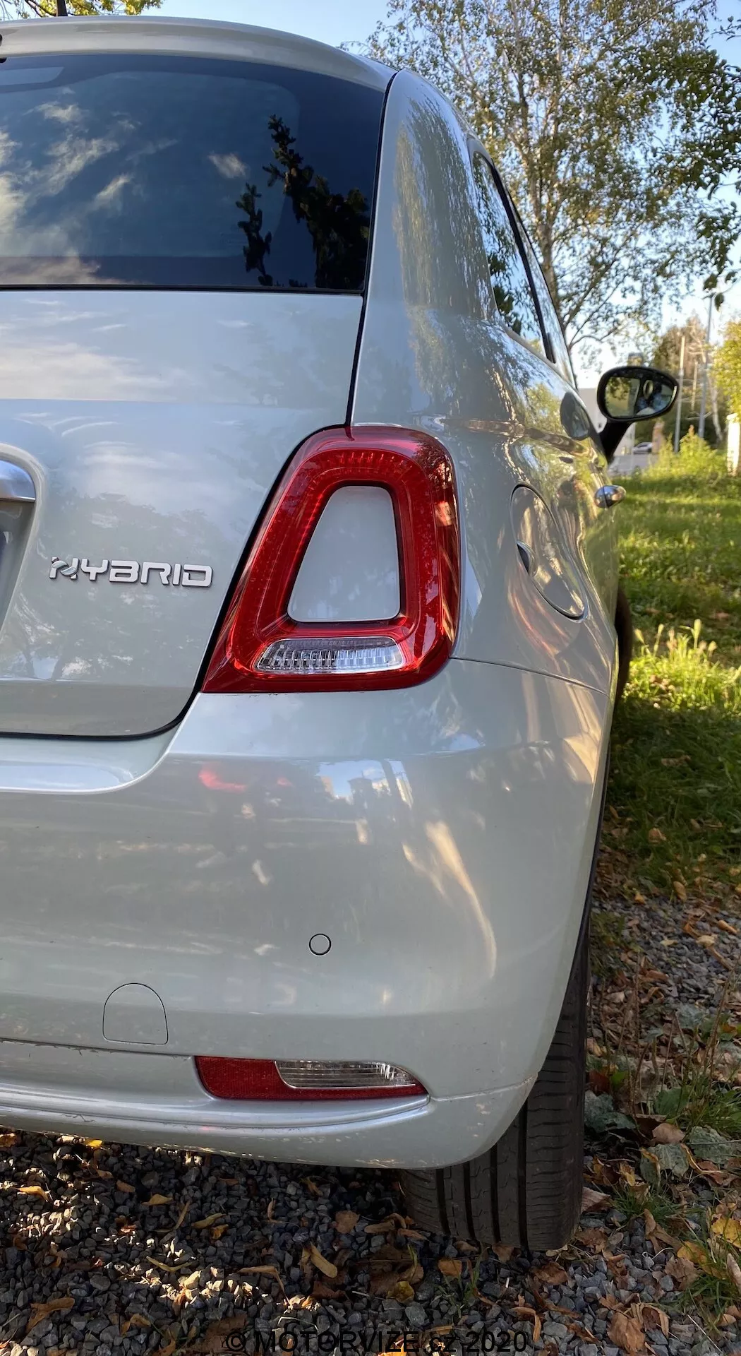 Widok tylno-prawy hybrydowego Fiata 500 z 2015 roku w metalicznym srebrze, pokazujący tylną lampę LED, odznakę hybrydową, tylny zderzak, częściowe widok felgi aluminiowej, przyciemnione tylne okno i błyszczącą karoserię; sfotografowany w podmiejskim parku z zielenią i drzewami.