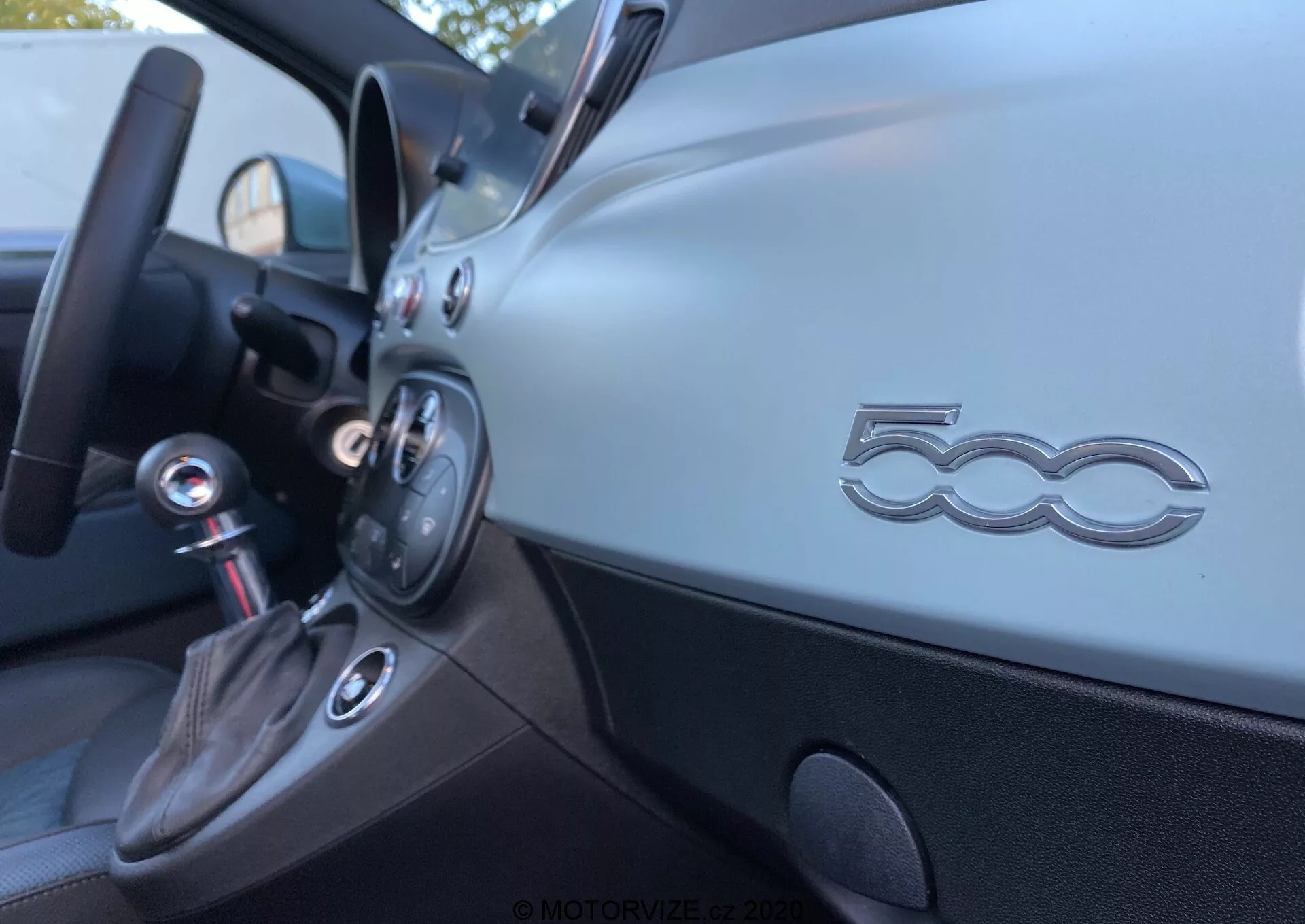 Widok wnętrza Fiata 500 2015 z przedniego siedzenia pasażera, ukazujący ukośny kąt na deskę rozdzielczą po stronie kierowcy. Matowa metaliczna deska rozdzielcza posiada wyraźne logo '500', przyciski kontrolne i okrągły nawiew. Poniżej znajduje się czarny panel i gałka zmiany biegów obszyta skórą z chromowanym wykończeniem, wskazująca na manualną skrzynię biegów. Wewnątrz drzwi znajduje się ergonomiczna klamka oraz schowek. Ostrość jest płytka, podkreślając emblemat '500', podczas gdy inne elementy są lekko rozmyte.