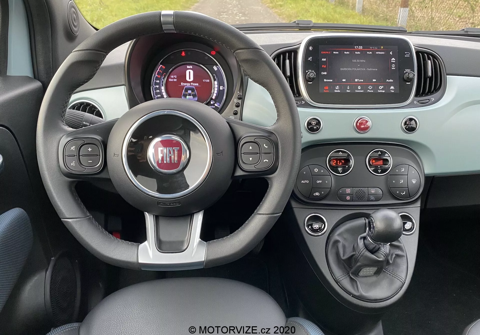 Widok wnętrza Fiata 500 2015 z perspektywy kierowcy pokazujący trójramienną kierownicę z przyciskami sterowania, częściowo widoczny cyfrowy zestaw wskaźników, wyłączony ekran dotykowy infotainment, przyciski i pokrętła klimatyzacji, manualną dźwignię zmiany biegów, dźwignię hamulca ręcznego oraz dwukolorowy schemat kolorystyczny podkreślający funkcjonalność i nowoczesne elementy projektowe.