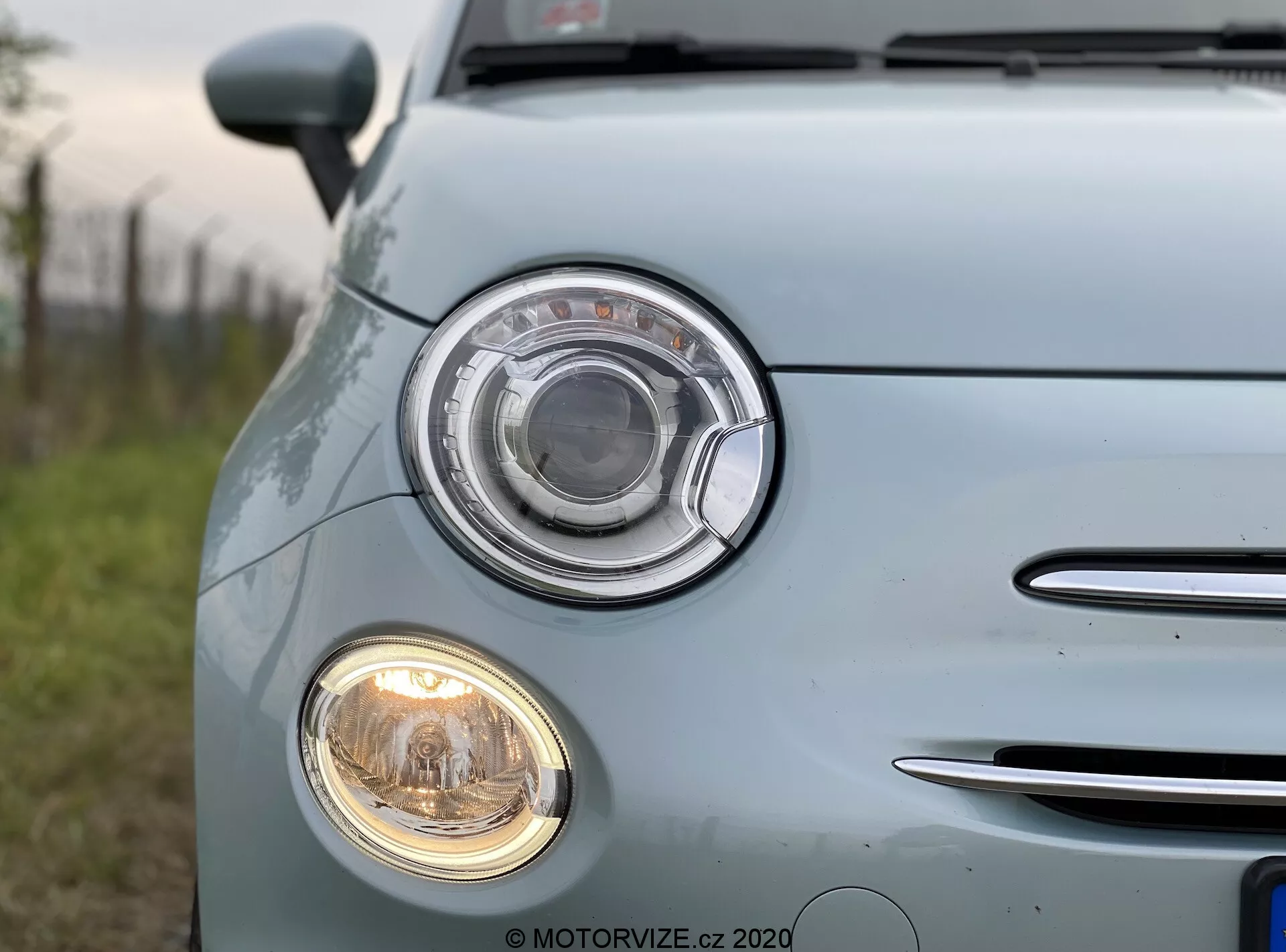 Zbliżenie na jasno-niebieski Fiat 500 z 2015 roku, uchwycony z przedniego lewego narożnika. Skupienie na charakterystycznym zespole reflektorów z okrągłym designem i przezroczystą soczewką, a także na mniejszym, okrągłym reflektorze przeciwmgielnym poniżej. Z lewej strony widoczny jest fragment osłony chłodnicy z poziomymi listewkami chromowanymi. Blada, niebieska, błyszcząca farba samochodu odbija naturalne światło, podkreślając dobrze utrzymaną powierzchnię. Znaki i logotypy pojazdu zostały rozmyte.