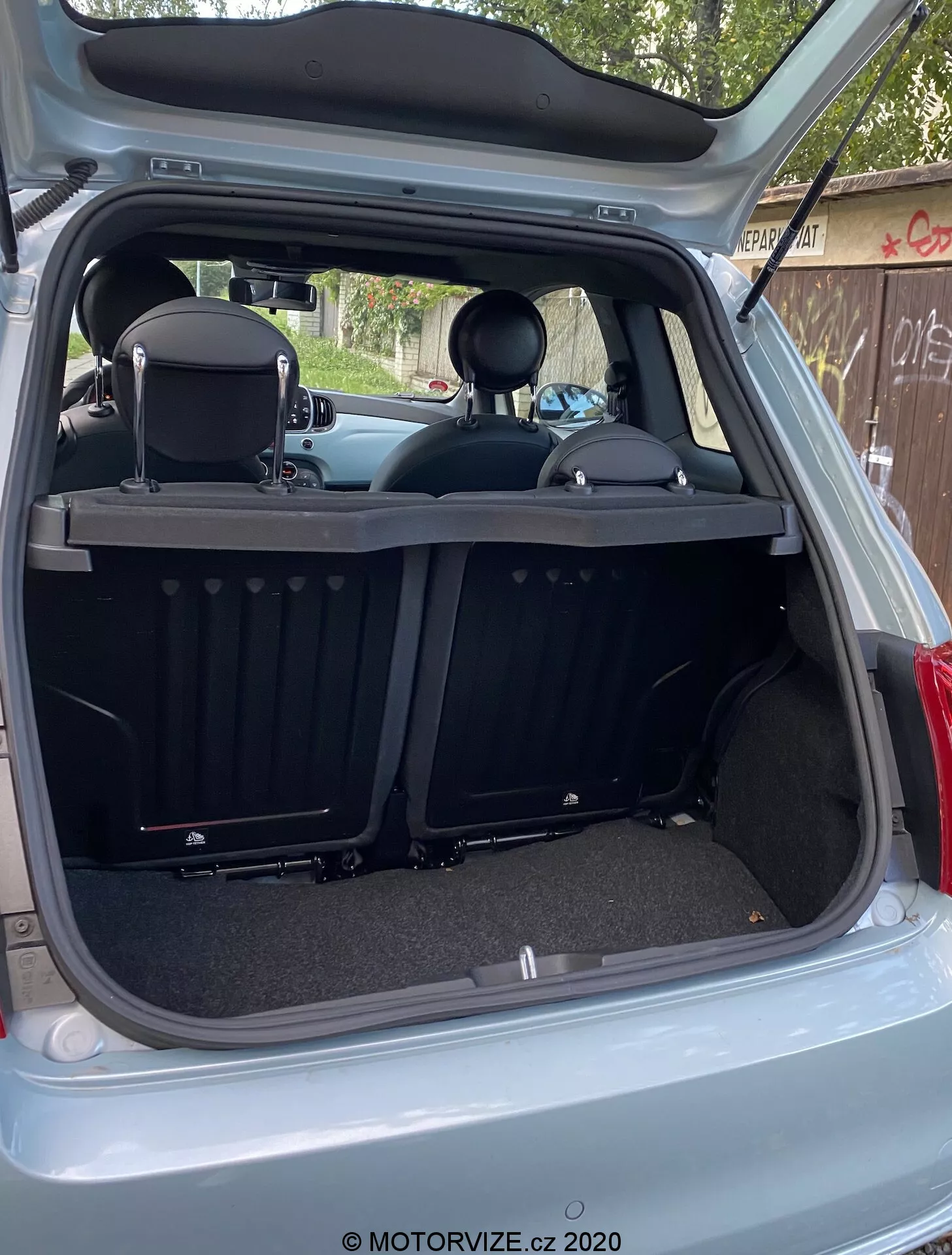 Zdjęcie pokazuje tylną część wnętrza Fiata 500 2015 z zewnętrznego tylnego kąta z całkowicie otwartymi drzwiami bagażnika. Przestrzeń bagażowa jest widoczna z nieco przesuniętego w prawo kąta, co ukazuje głębokość bagażnika i różne tylne elementy wnętrza. Nowoczesny, czysty design obejmuje wykładziny dywanowe, ryflowaną, zwijaną pokrywę bagażnika, składane tylne siedzenia z zagłówkami oraz listwę ochronną. Cechy technologiczne obejmują wewnętrzne światło, gniazdo 12V, plastikowe listwy, okrągłe wycięcia oraz boczne panele.