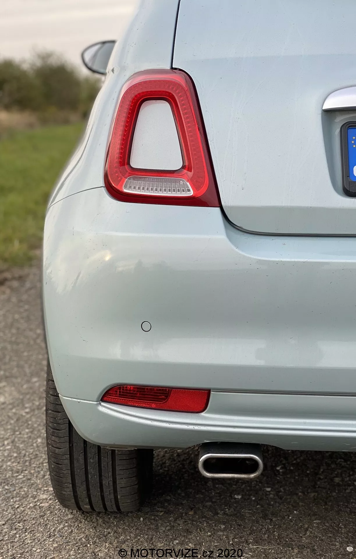 Zbliżenie z ukosa tylnej lewej strony jasnego samochodu, z podkreśleniem charakterystycznej tylnej lampy z przezroczystym czerwonym kloszem i technologią LED, czerwonego odblasku, montowanego na równo czujnika parkowania i rury wydechowej z chromowanym wykończeniem. Jasna metaliczna farba i płytka głębia ostrości podkreślają czysty i dobrze utrzymany stan pojazdu.