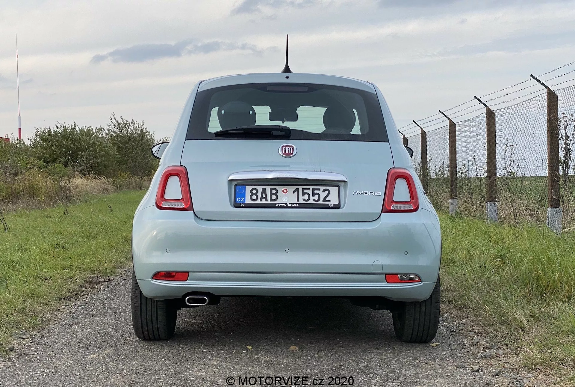 Tylny widok na jasno niebieski samochód Fiat 500 2015. Ujęcie na wysokości oczu uchwyca stojący pojazd, ukazując cały jego tył, w tym klapę bagażnika, tylną szybę, tylne światła, tylny zderzak oraz tablicę rejestracyjną. Cecha to widoczne logo producenta, tylna wycieraczka, wysoko umieszczone światło stopu i okrągłe tylne światła z dodatkowymi segmentami. Tablica rejestracyjna zamontowana jest na środku klapy bagażnika, pod emblematem i nad płaskim tylnym zderzakiem z wbudowanymi odbłyśnikami. Krótka antena jest umieszczona na środku dachu bliżej tyłu. Zachmurzone niebo i wiejski krajobraz, z płotem po prawej stronie i trawą poniżej.