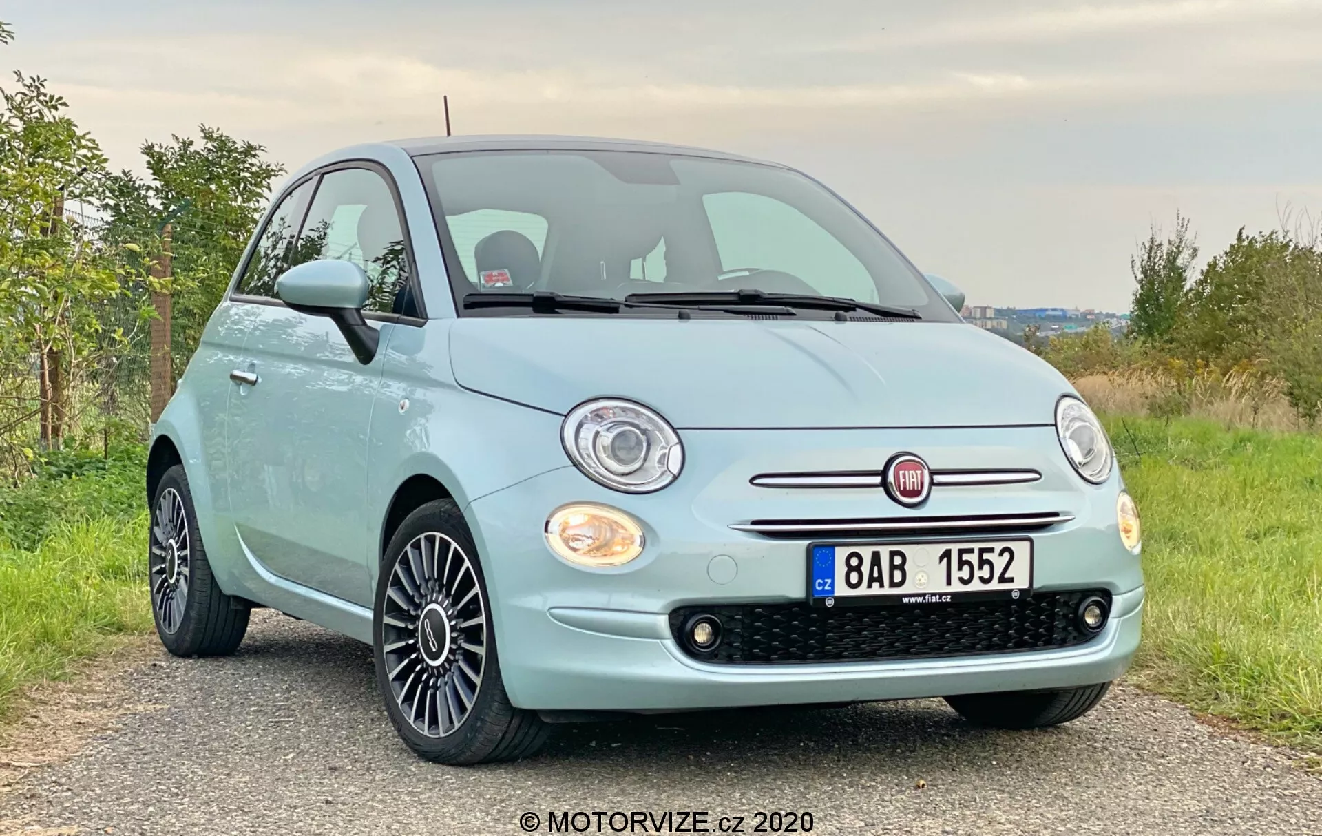 Widok przedni pod kątem trzech czwartych błękitnego Fiata 500 z 2015 roku z chromowaną kratką i okrągłymi reflektorami halogenowymi pod nieco niskim kątem. Obrazek podkreśla przednią fasadę, kratkę, reflektory, maskę i aluminiowe koła z czarnym wieloszprychowym wzorem. Samochód jest zaparkowany na żwirowej lub ziemnej drodze na obszarze wiejskim z zieloną roślinnością w tle.