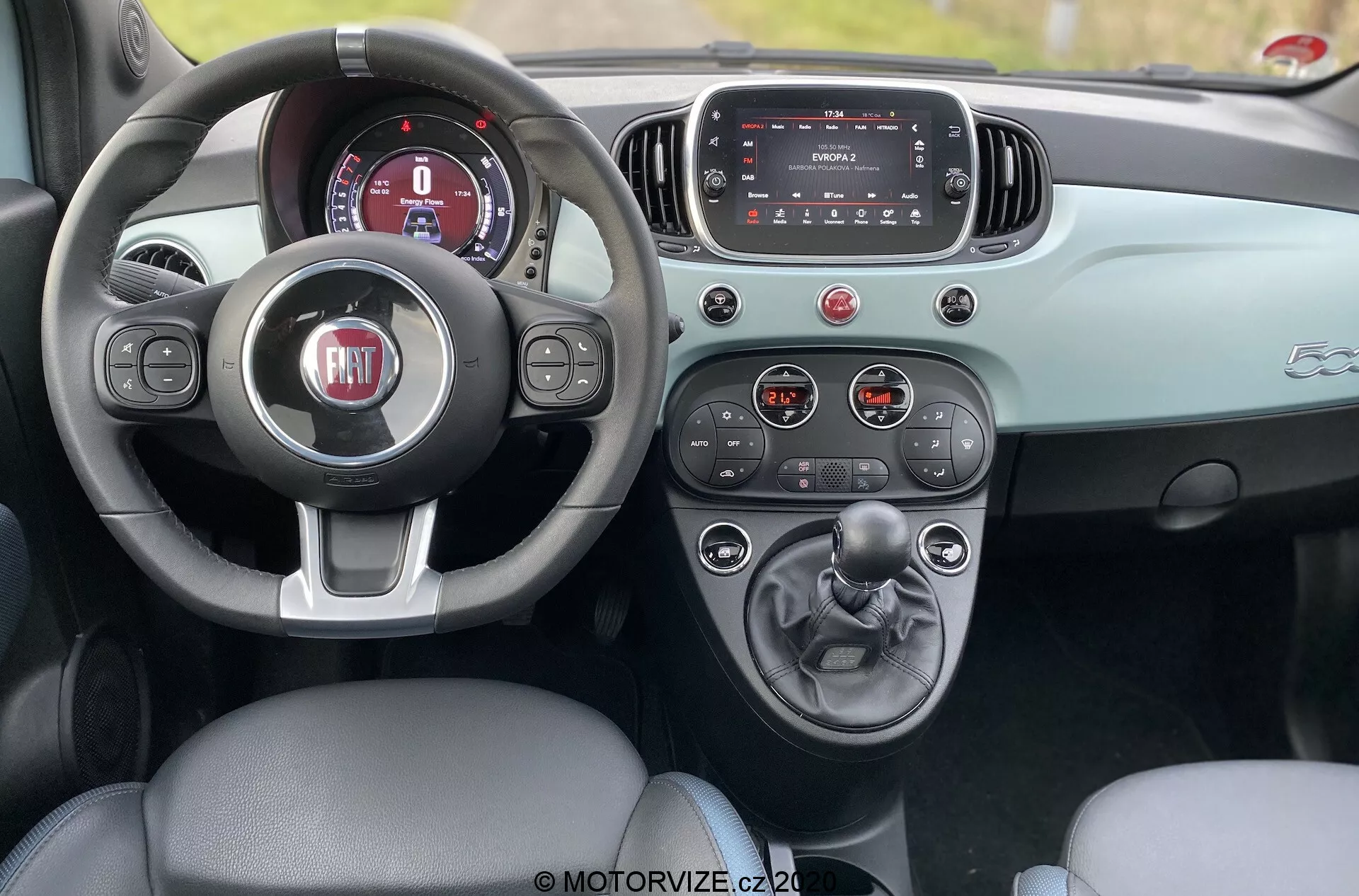 Zdjęcie wnętrza Fiata 500 z 2015 roku widziane z perspektywy tylnej kanapy, ukazujące przedni kokpit z skórzanym siedzeniem kierowcy, trójramienną kierownicą z logo FIAT, cyfrowym panelem instrumentów, ekranem dotykowym systemu infotainment i konsolą środkową z manualną skrzynią biegów i uchwytami na kubki. Deska rozdzielcza i drzwi mają dwukolorową tonację, która podkreśla estetyczny design. Zdjęcie wykonane w ciągu dnia, podkreślające wyrafinowane wnętrze i funkcjonalność samochodu.