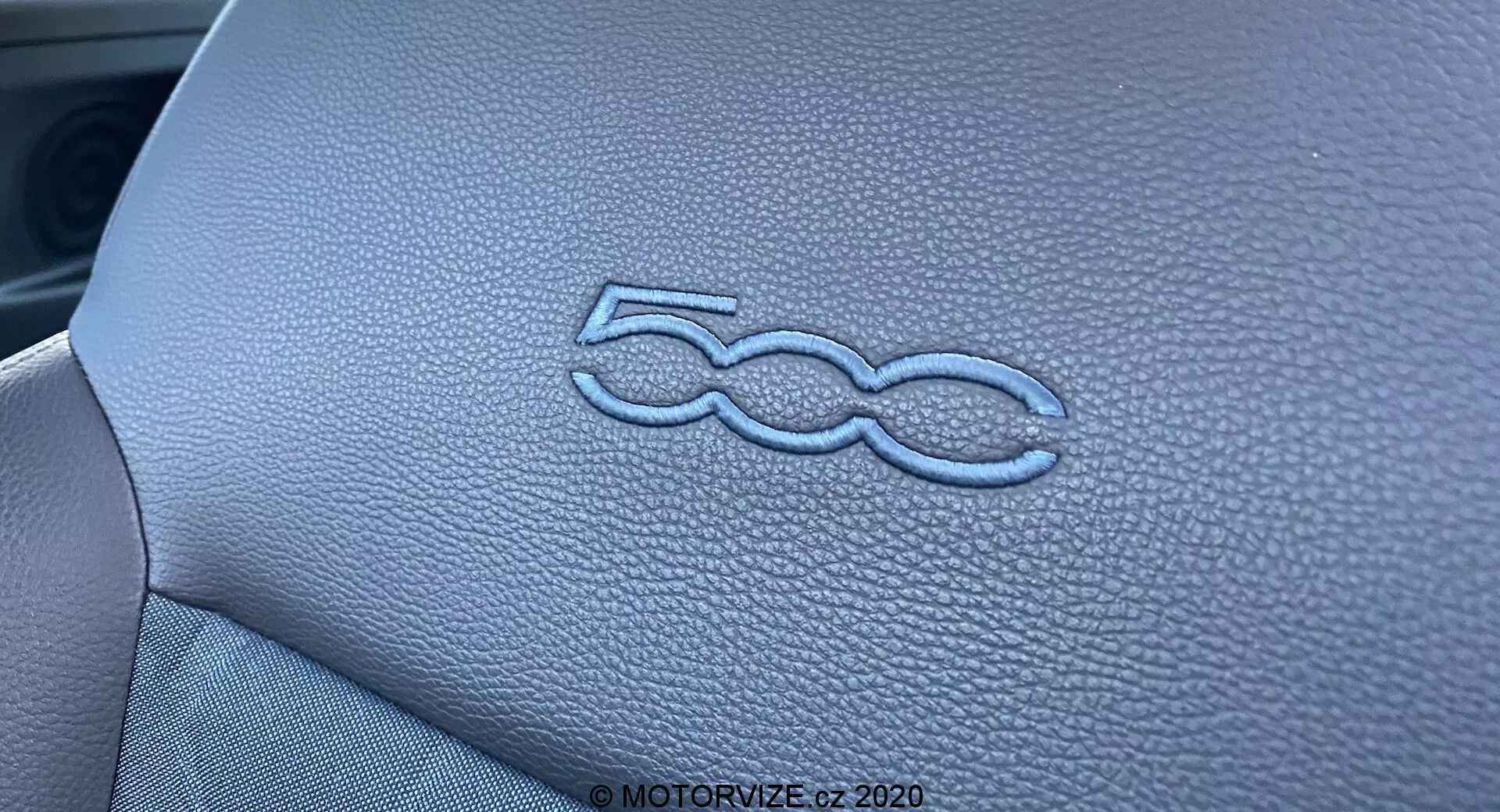 Zbliżenie ukazujące stylizowane, wytłaczane logo '500' na desce rozdzielczej wnętrza Fiata 500 2015. Skórzana tekstura i precyzyjne szwy panelu są podkreślone przez oświetlenie i wąską głębię ostrości, podkreślając wysoką jakość wykonania wnętrza pojazdu.