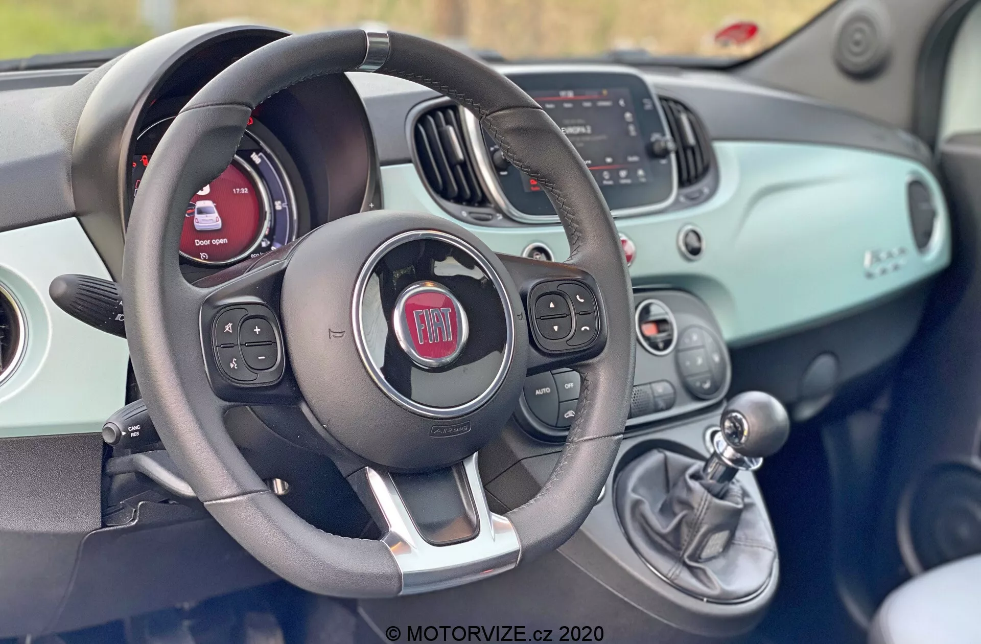 Widok wnętrza Fiata 500 z roku 2015 z miejsca kierowcy, pokazujący kierownicę z wbudowanymi sterowaniami audio, poleceniami głosowymi i prawdopodobnie tempomatem. Za kierownicą widać zestaw wskaźników z cyfrowym ekranem i tradycyjnymi wskaźnikami. Deska rozdzielcza ma powierzchnię w kolorze nadwozia i zawiera ekran infotainment wyświetlający informacje o radiu i mediach oraz ręczne sterowanie HVAC. Po prawej stronie kierownicy znajduje się dźwignia zmiany biegów, co wskazuje na manualną skrzynię biegów. Cały design jest nowoczesny, z sportowym, dwukolorowym wyglądem.
