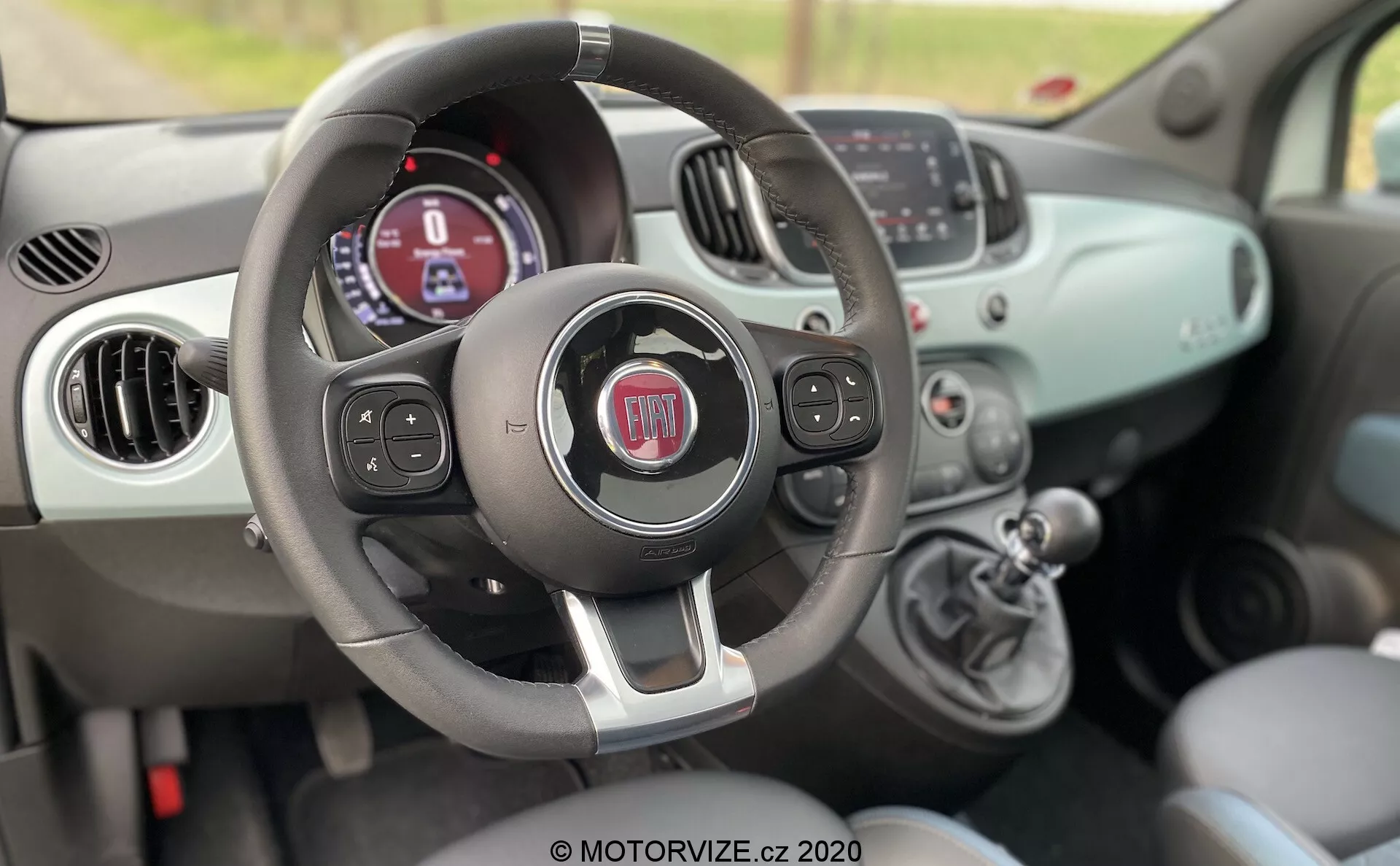 Widok wnętrza Fiata 500 z 2015 roku z perspektywy pasażera, ukazujący kierownicę z przyciskami wielofunkcyjnymi, zestaw wskaźników z wyświetlaczem cyfrowym, ręczne sterowanie klimatyzacją i gałkę zmiany biegów.