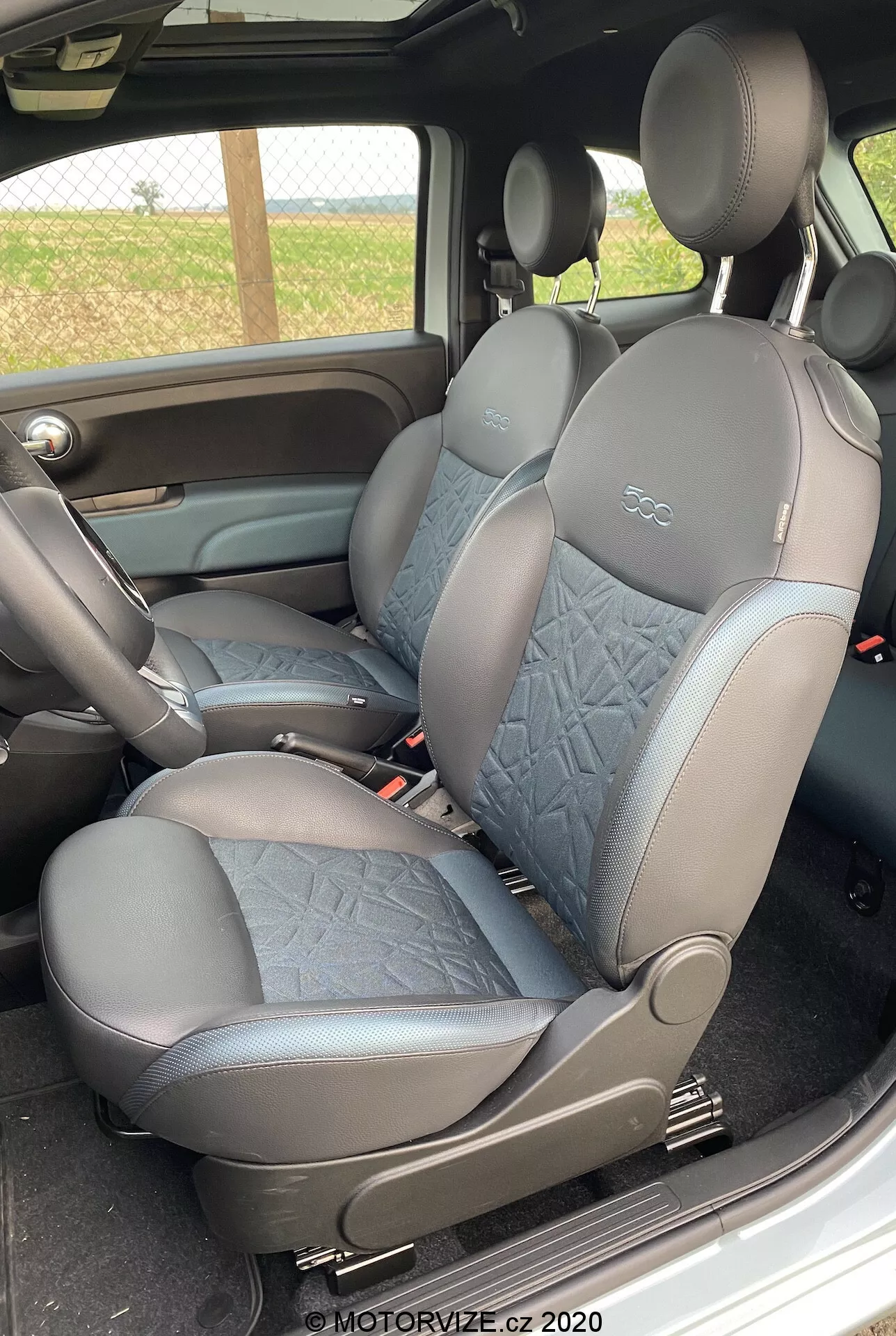 Zdjęcie przedstawia wnętrze Fiata 500 z 2015 roku, miejsce pasażera, widok boczny przednich siedzeń. Tapicerka ze skóry i pikowanej tkaniny. Zintegrowane zagłówki. Ciemny kolor dla praktyczności. Widoczna dźwignia ręcznej regulacji siedzenia. Częściowo widoczny podłokietnik środkowej konsoli. Spójność kolorów i materiałów z panelem drzwi. Znak wodny z 2020 roku.