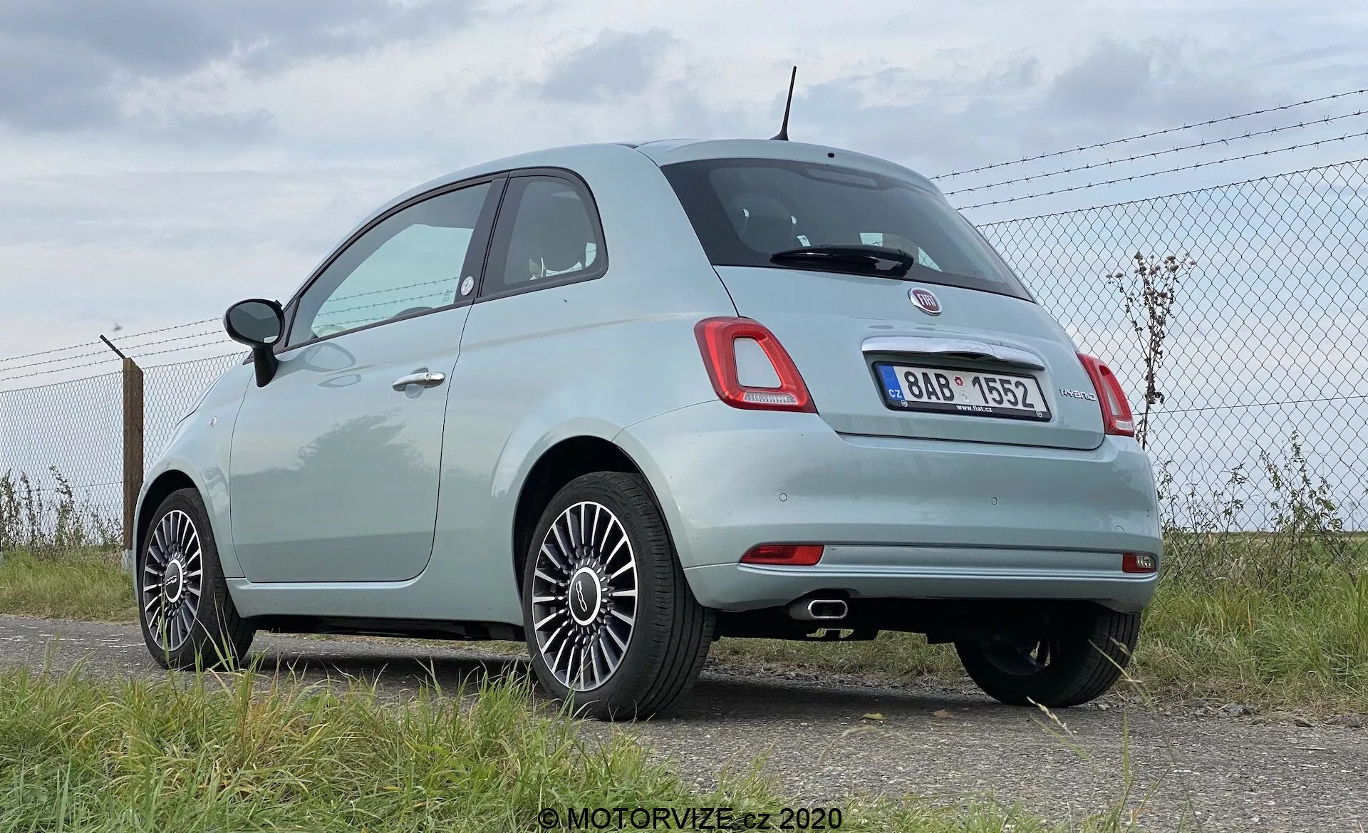 Trzyczwarte tylne ujęcie jasnego nowoczesnego hatchbacka Fiat 500, ukazujące charakterystyczne zaokrąglone tylne światła oraz centralny chromowany znaczek nad tylną tablicą rejestracyjną. Obraz uwydatnia zaokrągloną linię dachu pojazdu, chromowane listwy wokół bocznych okien oraz wieloramienne felgi aluminiowe. Jasny, pastelowy lakier samochodu wskazuje na jego stosunkowo nowy lub dobrze utrzymany stan. Pojazd jest zaparkowany w pobliżu ogrodzenia z łańcuszka na utwardzonej powierzchni, z widocznym na tle niebem i horyzontem, co sugeruje lotnisko lub otwartą przestrzeń. Możliwe funkcje technologiczne to tylne czujniki parkowania zintegrowane z czarną sekcją tylnego zderzaka.