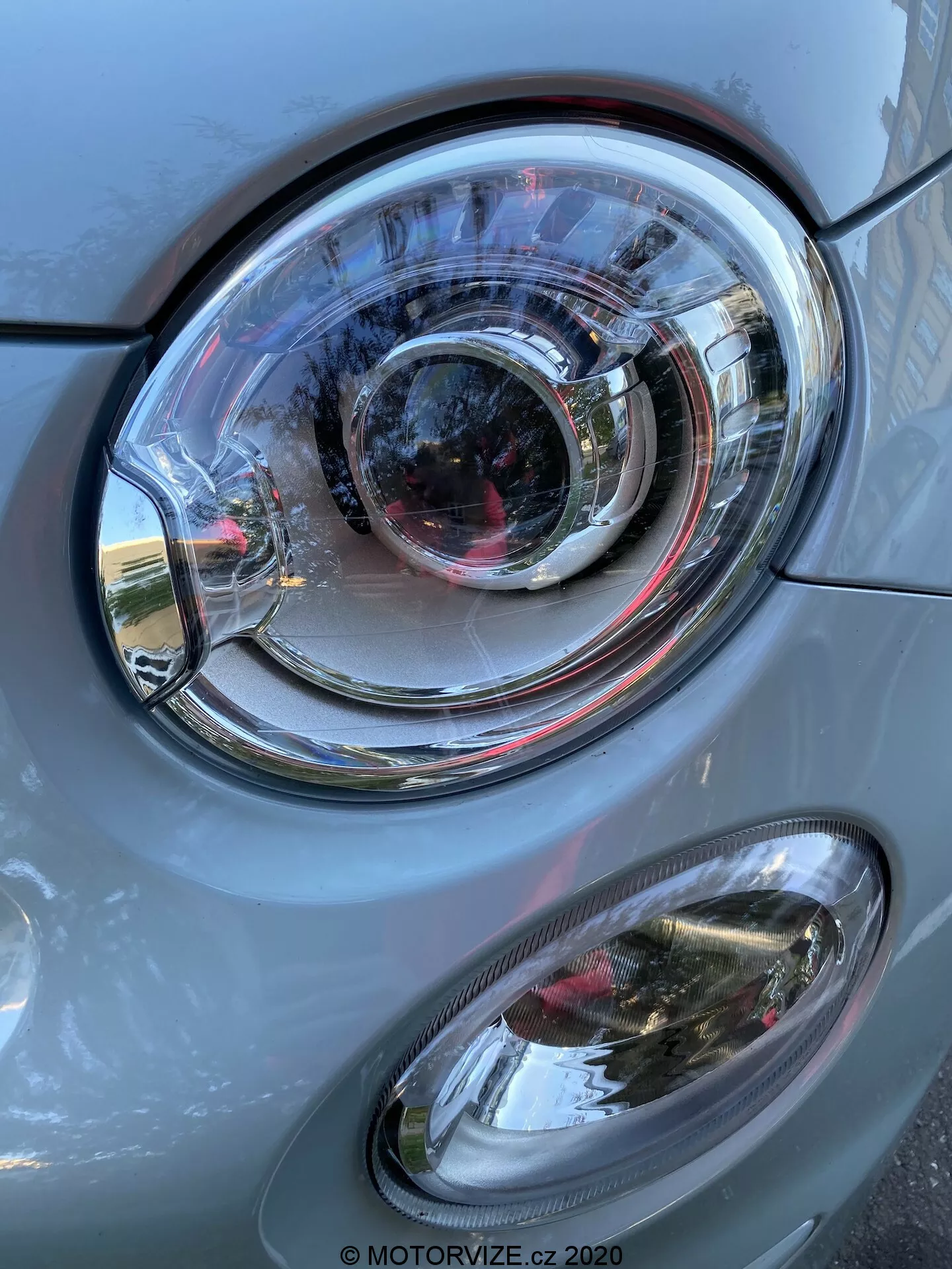 Zbliżenie przedniej lewej strony samochodu Fiat 500 2015, skupiające się na zespole reflektorów. Obraz w pionowej orientacji przedstawia wyraźny główny reflektor z przezroczystą soczewką z poliwęglanu o skomplikowanej konstrukcji z koncentrycznymi pierścieniami, co wskazuje na styl projektora i być może adaptacyjną technologię oświetlenia. Poniżej głównego reflektora znajduje się owalny, drugorzędny reflektor przeciwmgielny z przezroczystą osłoną. Panele karoserii samochodu w kolorze szarym z metalicznym połyskiem mają gładkie wykończenie, odbijając światło dzienne i elementy otoczenia, takie jak budynki i niebo. Zespół reflektorów i reflektor przeciwmgielny są w dobrym stanie, co wskazuje na dobrze utrzymany lub nowy pojazd.