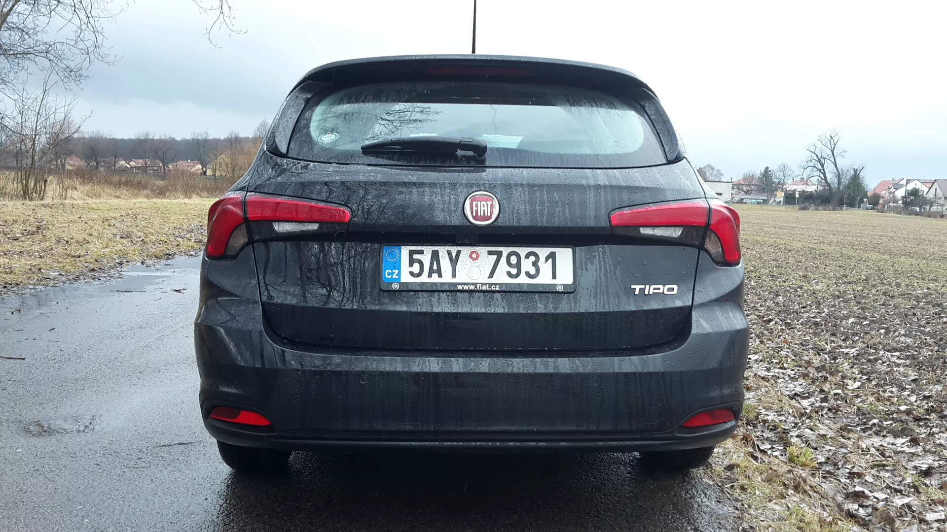 Tylny widok sedana Fiat Tipo 2016 pod pochmurnym niebem, z identyfikacją marki i modelu na bagażniku, szczegółowymi światłami tylnymi i trzecim światłem stopu, tablicą rejestracyjną i widokiem na wiejską okolicę w tle